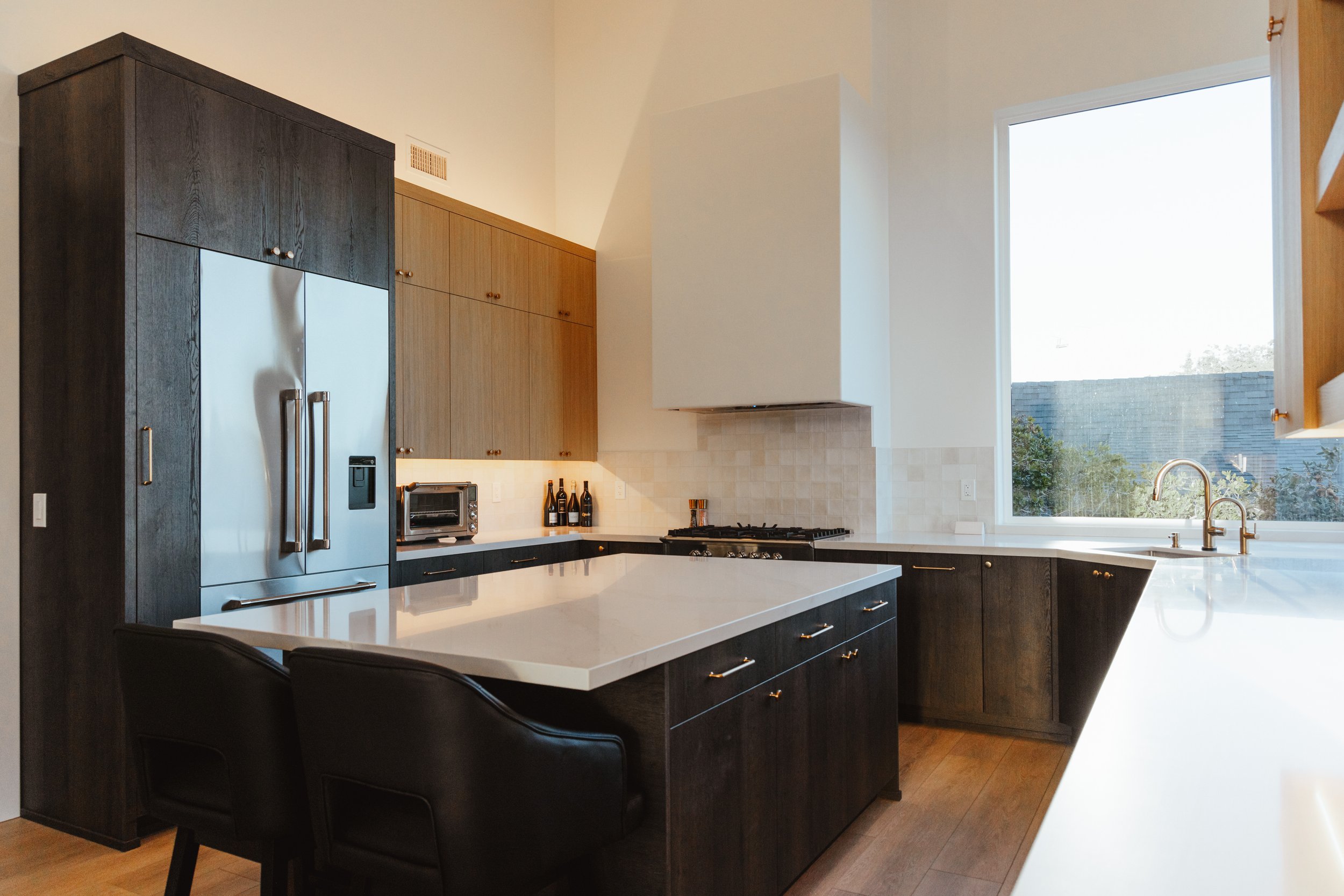 Modern kitchen with black and wood cabinetry, white countertops, stainless steel refrigerator, and large window overlooking outdoors.