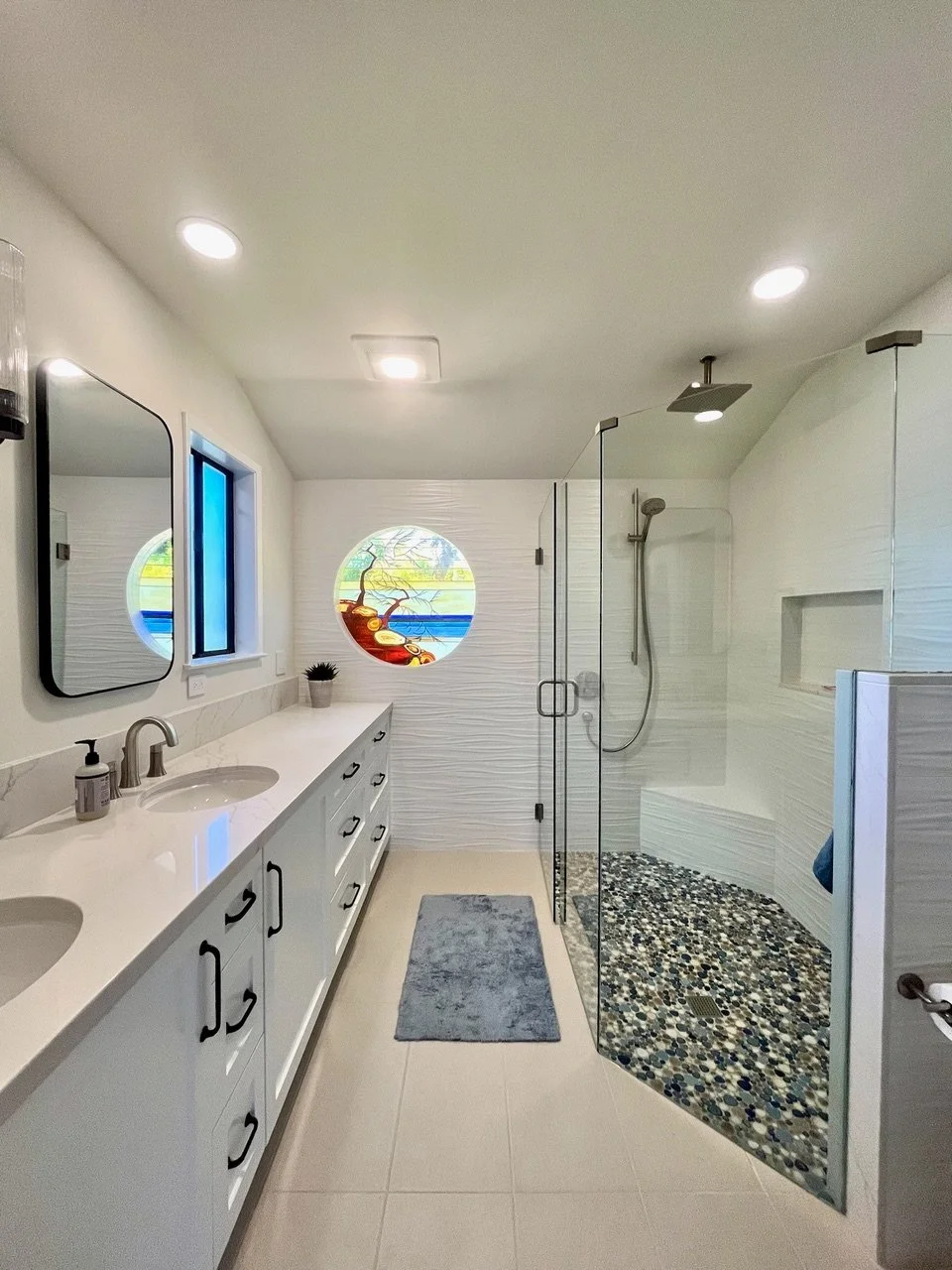 Modern white bathroom with dual sinks, black handles, large mirror, colorful round window, glass-enclosed shower with pebble floor and built-in shelf, gray bath mat.