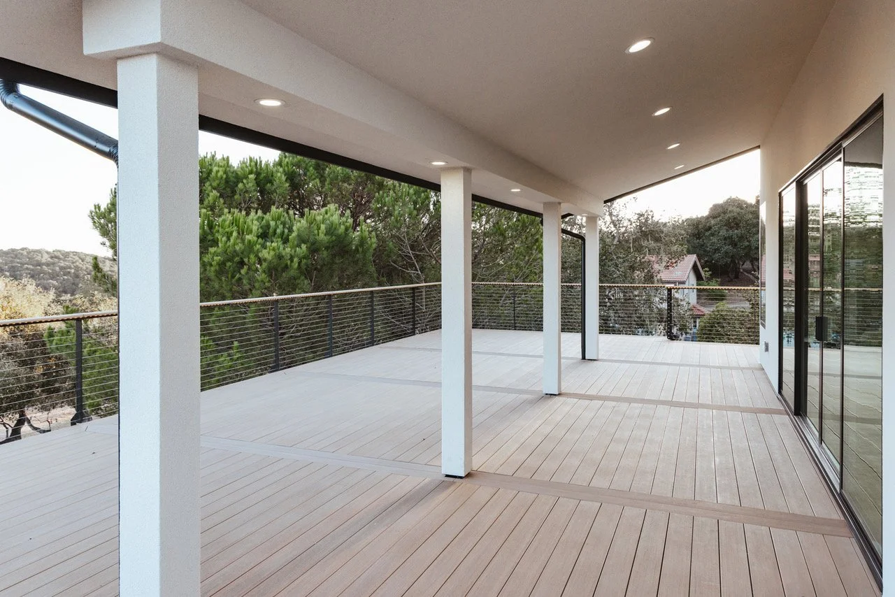 Empty modern balcony or patio with wooden flooring, white columns, sliding glass doors, and a metal railing, overlooking trees and a neighboring house.