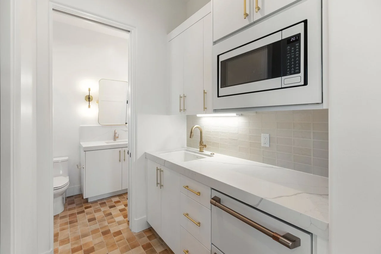 A small modern kitchen with white cabinets, a white marble countertop, and a built-in microwave. There are gold handles on the cabinets, a gold faucet, and a beige tile backsplash. In the back, visible through an open door, there is a white bathroom with a toilet, a small white sink with gold fixtures, and a wall-mounted light fixture.