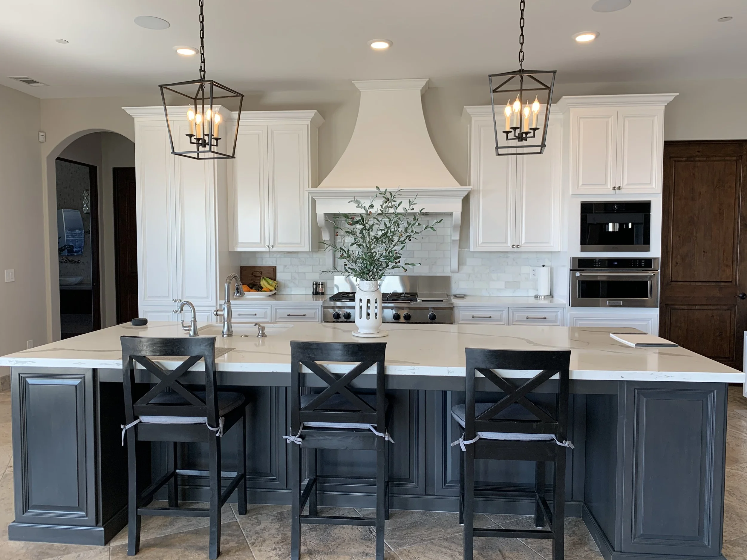 Modern kitchen with white cabinets, a central island with dark base and white countertop, black chairs with tied cushions, a large potted plant on the island, stainless steel appliances, two black and metal chandeliers hanging from the ceiling, and n