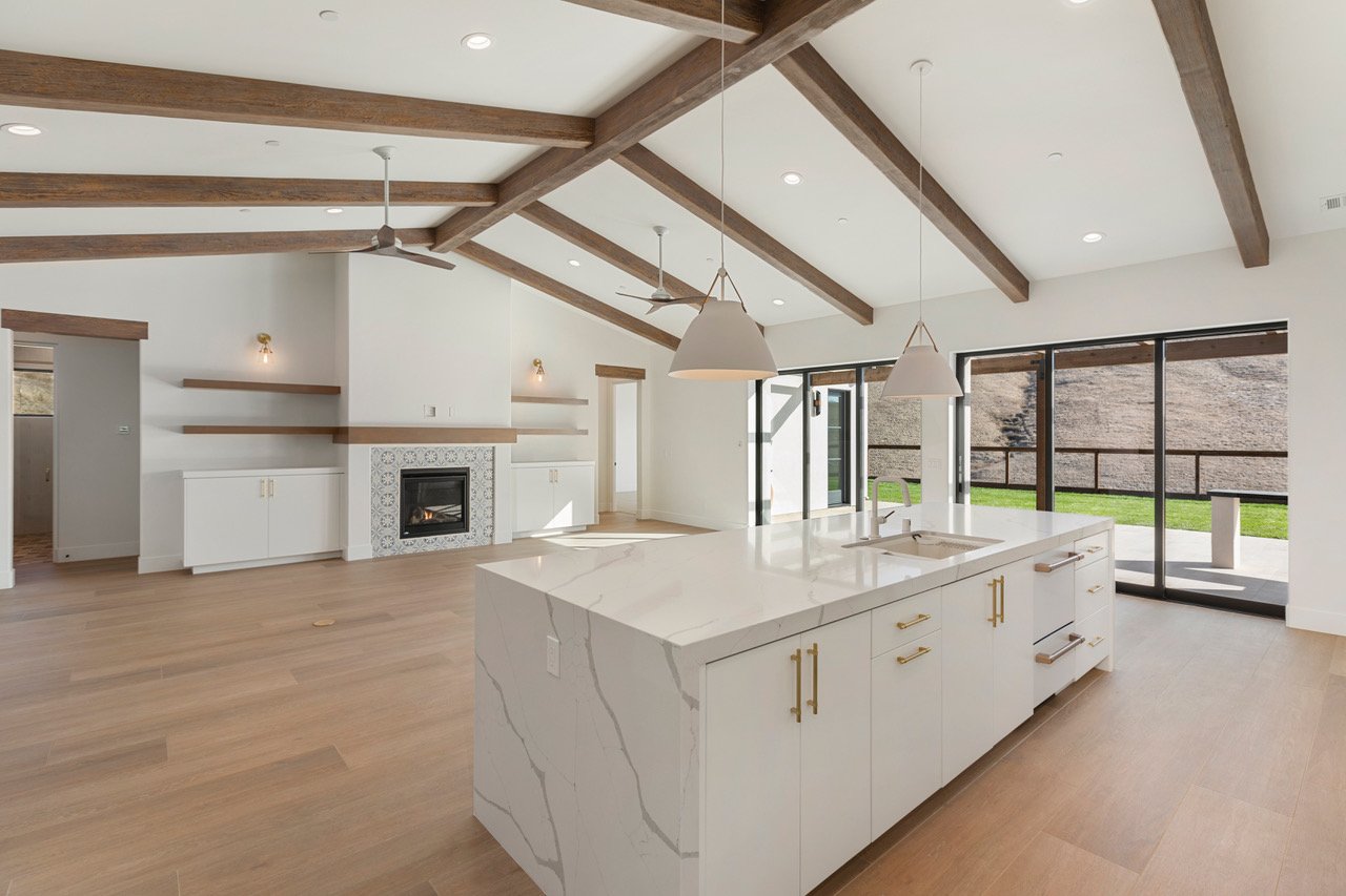 Modern open-concept living space with a kitchen island, white cabinets, gold hardware, large windows, a fireplace, and a vaulted ceiling with wooden beams.