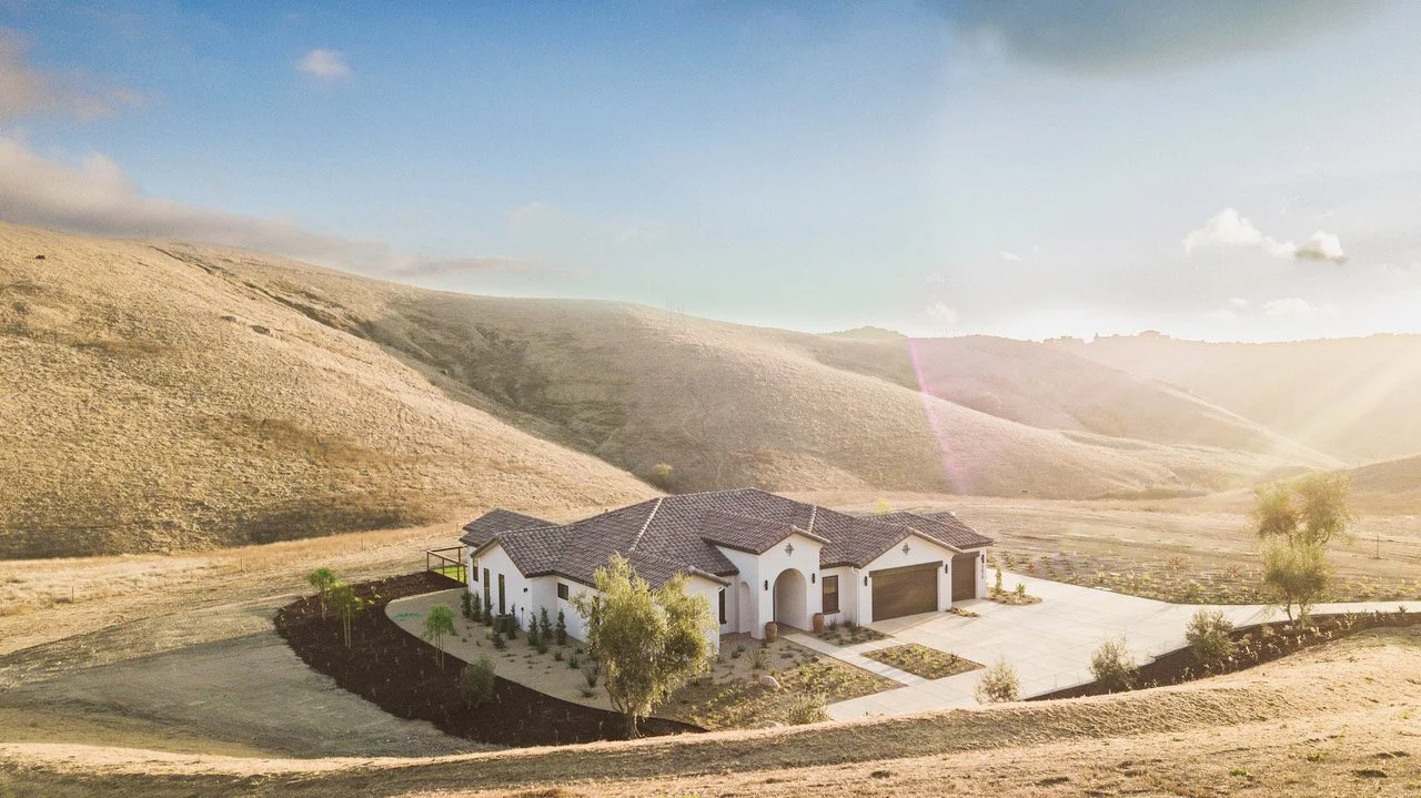 A white house with a tiled roof, surrounded by a landscaped yard with trees, set in a hilly, semi-arid landscape under a partly cloudy sky with sunlight.