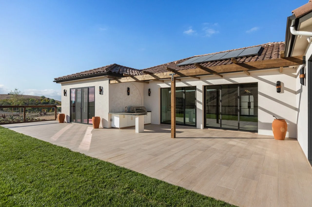 Backyard patio of a house with sliding glass doors, outdoor kitchen, large terracotta planters, and solar panels on the roof under a clear blue sky.