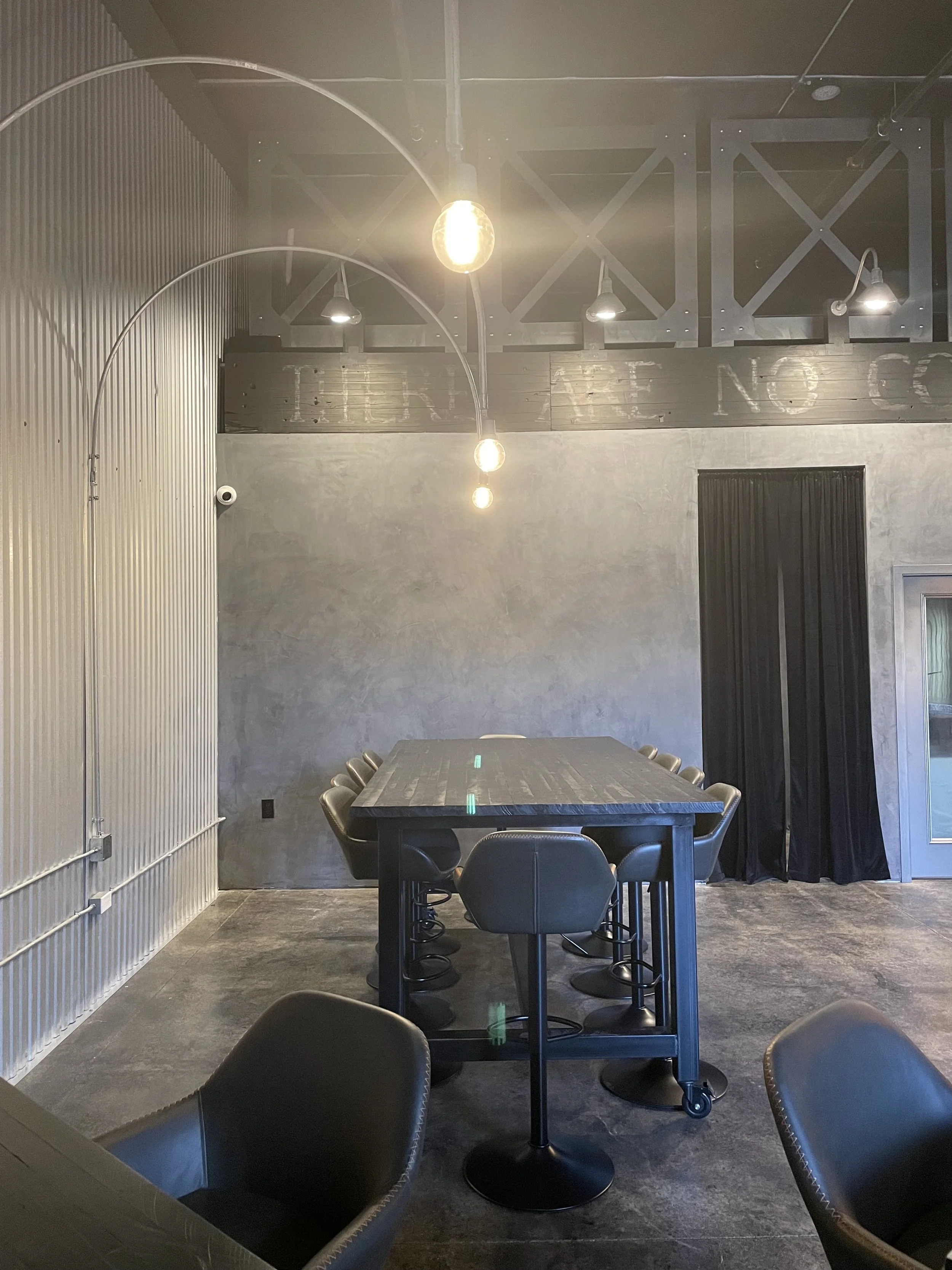 Indoor dining area with a long black table surrounded by black chairs, exposed lighting fixtures hanging from the ceiling, and a wall with black curtains and a door.