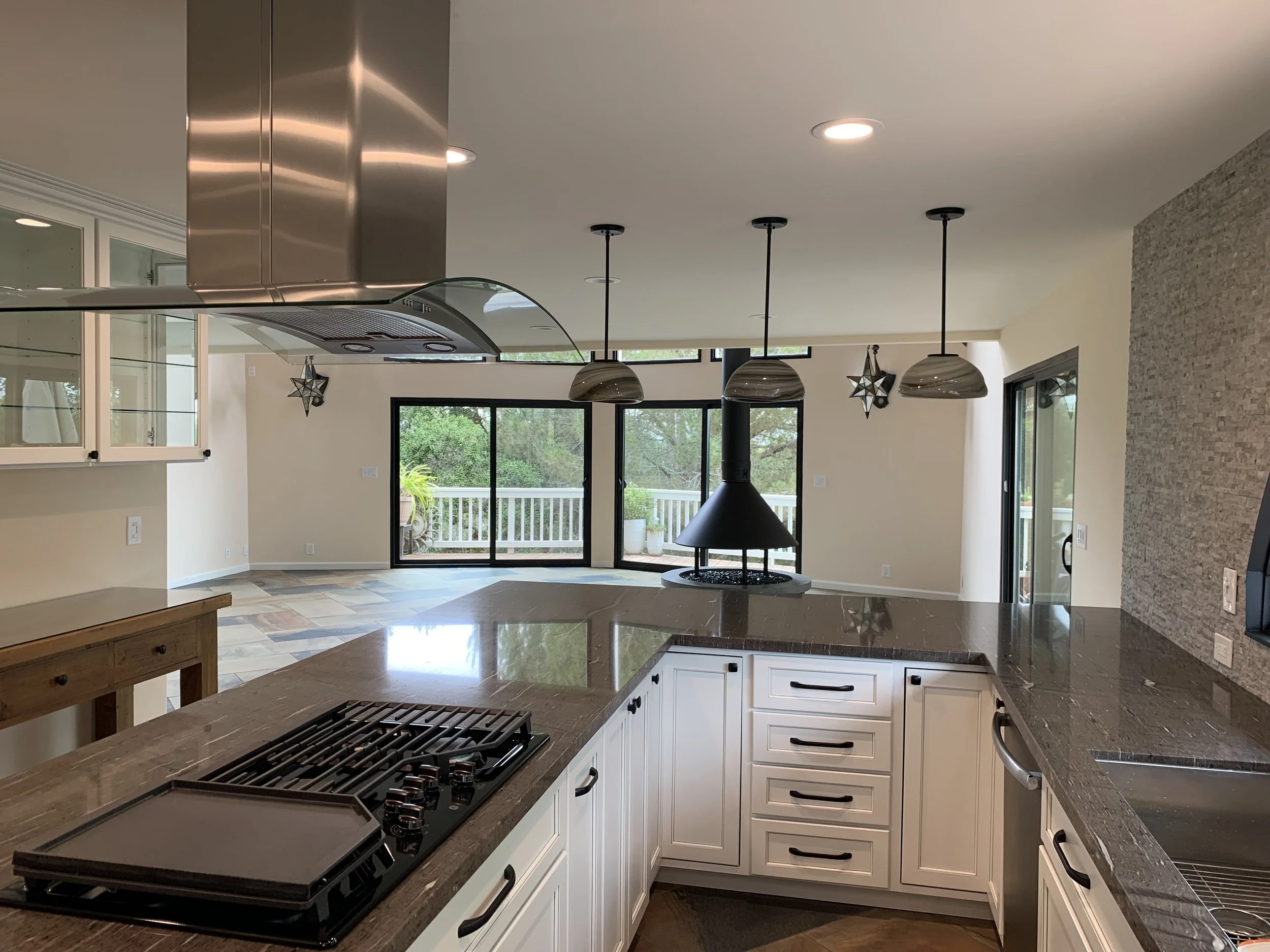Modern kitchen with white cabinets, dark granite countertops, a gas stove, and pendant lights hanging from the ceiling. Large windows and sliding glass doors reveal a view of outdoor greenery and a deck.