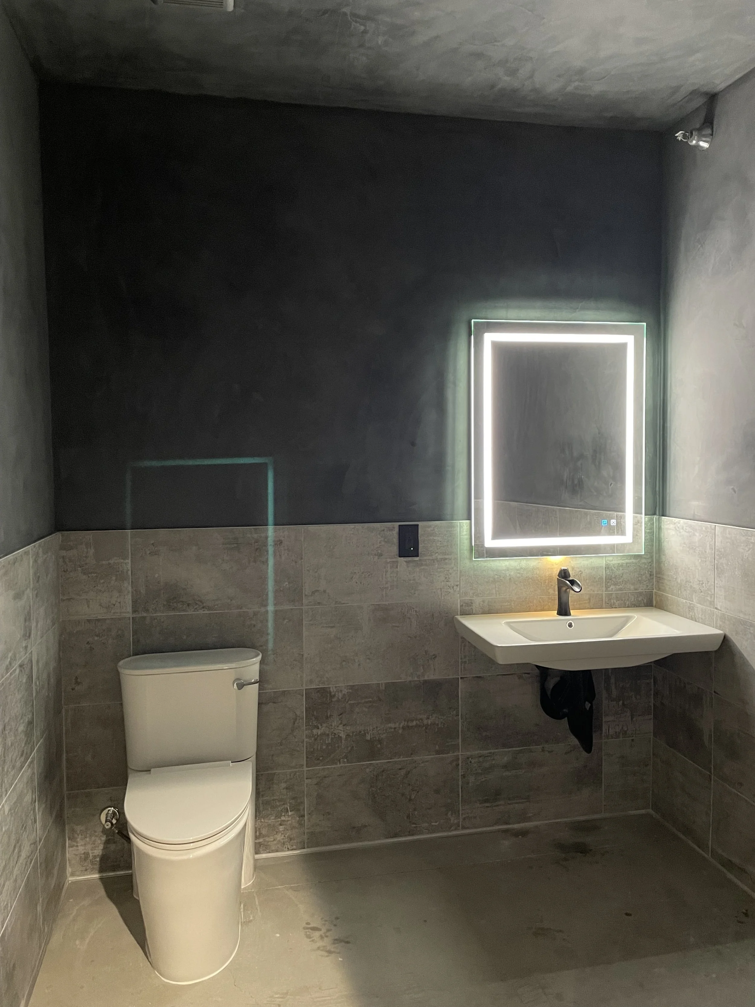 A modern bathroom with dark tiled walls, a white toilet, a white sink with a black faucet, and a rectangular mirror illuminated with LED lights.