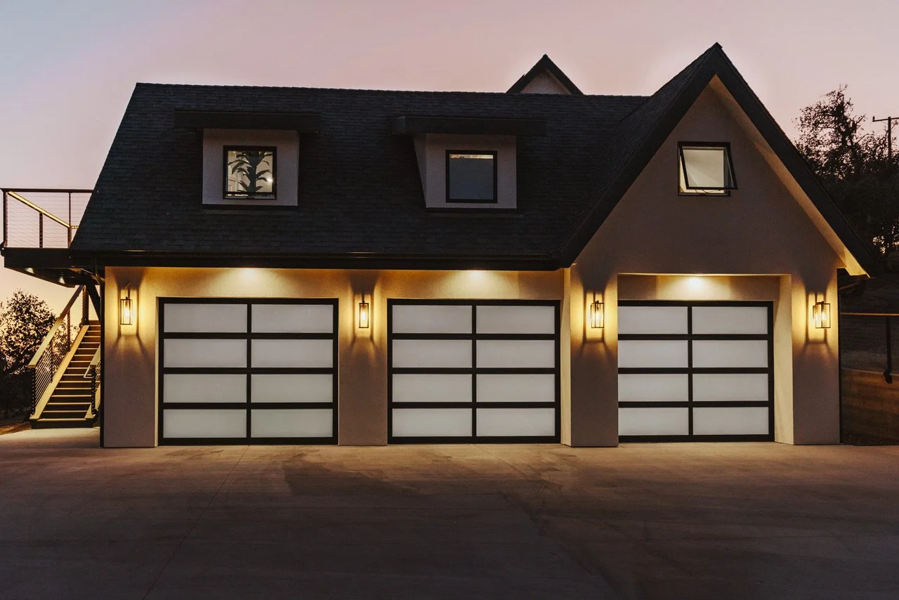 A modern house with a triple garage, lit up at dusk, with black-framed garage doors, exterior lights, and a staircase leading to a side balcony.