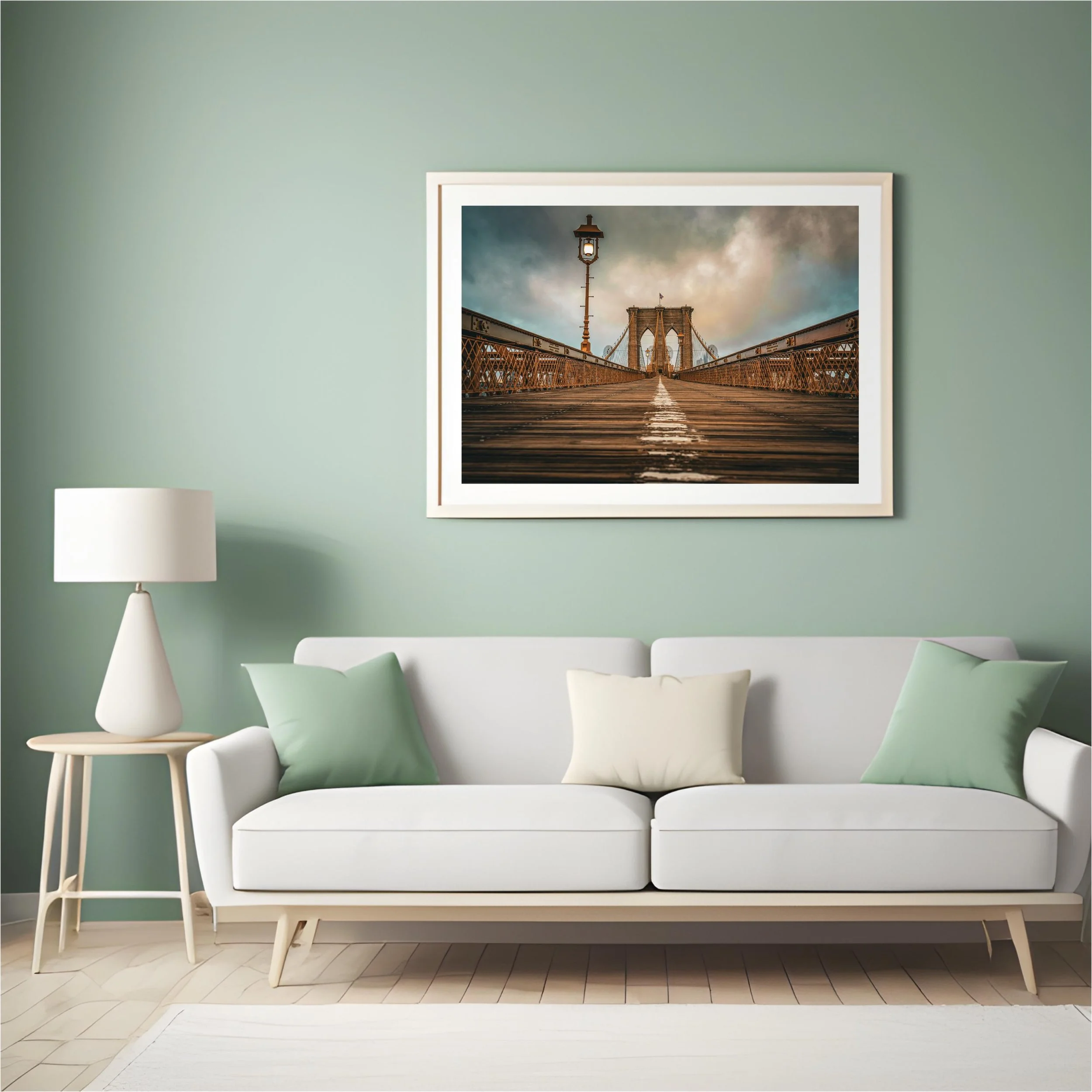 Framed photograph of Brooklyn Bridge viewed from below, featuring a wooden walkway, street lamp, and cloudy sky, hung on a green wall above a white sofa with green and beige pillows in a living room.
