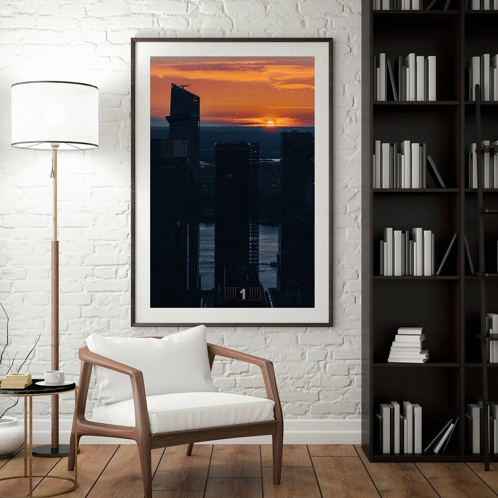 Interior of a modern living room with a white brick wall, a framed photo of a city skyline at sunset, a white armchair, a small side table with a cup and book, a tall floor lamp, and a black bookshelf filled with white and black books.