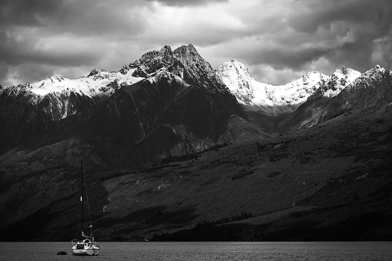 Black and white landscape of a mountain range with snow-capped peaks, a dark cloudy sky, and a sailboat on calm water in the foreground.