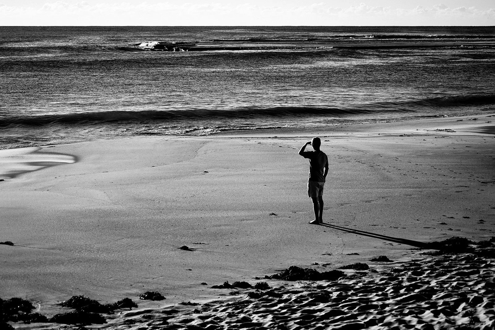 A person standing on a beach, looking out at the ocean, with one hand raised to their forehead in a saluting or shielding gesture, captured in black and white.