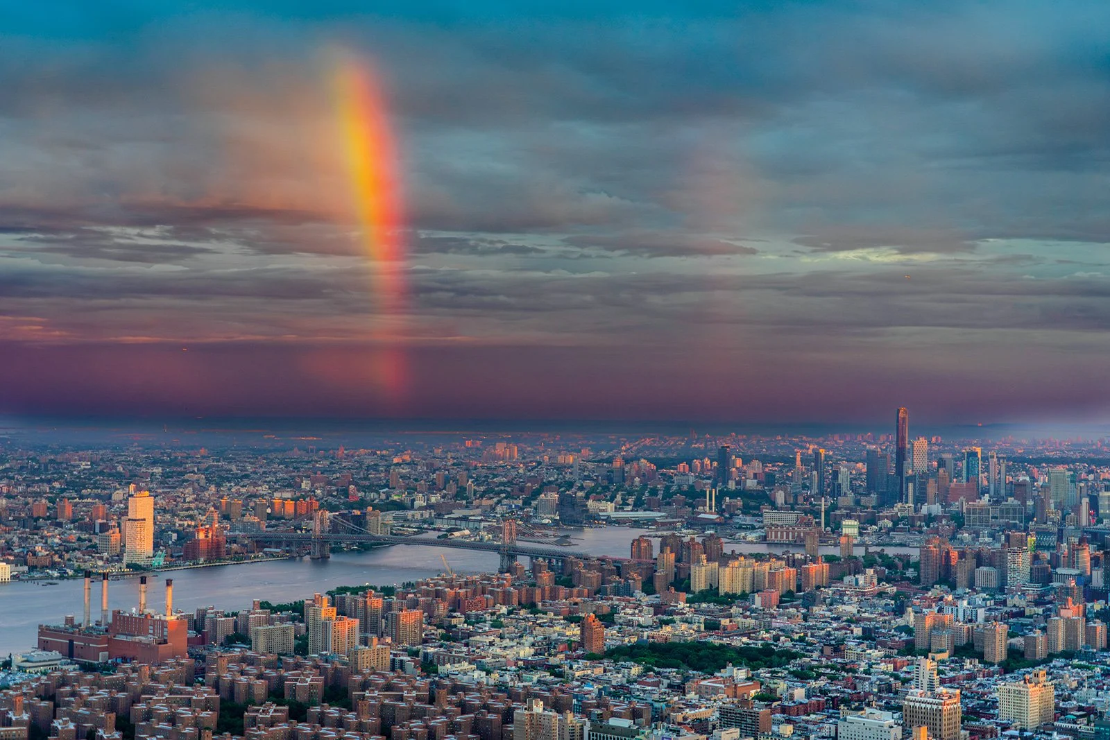 A city skyline at sunset with a rainbow in the cloudy sky over a river and various buildings.
