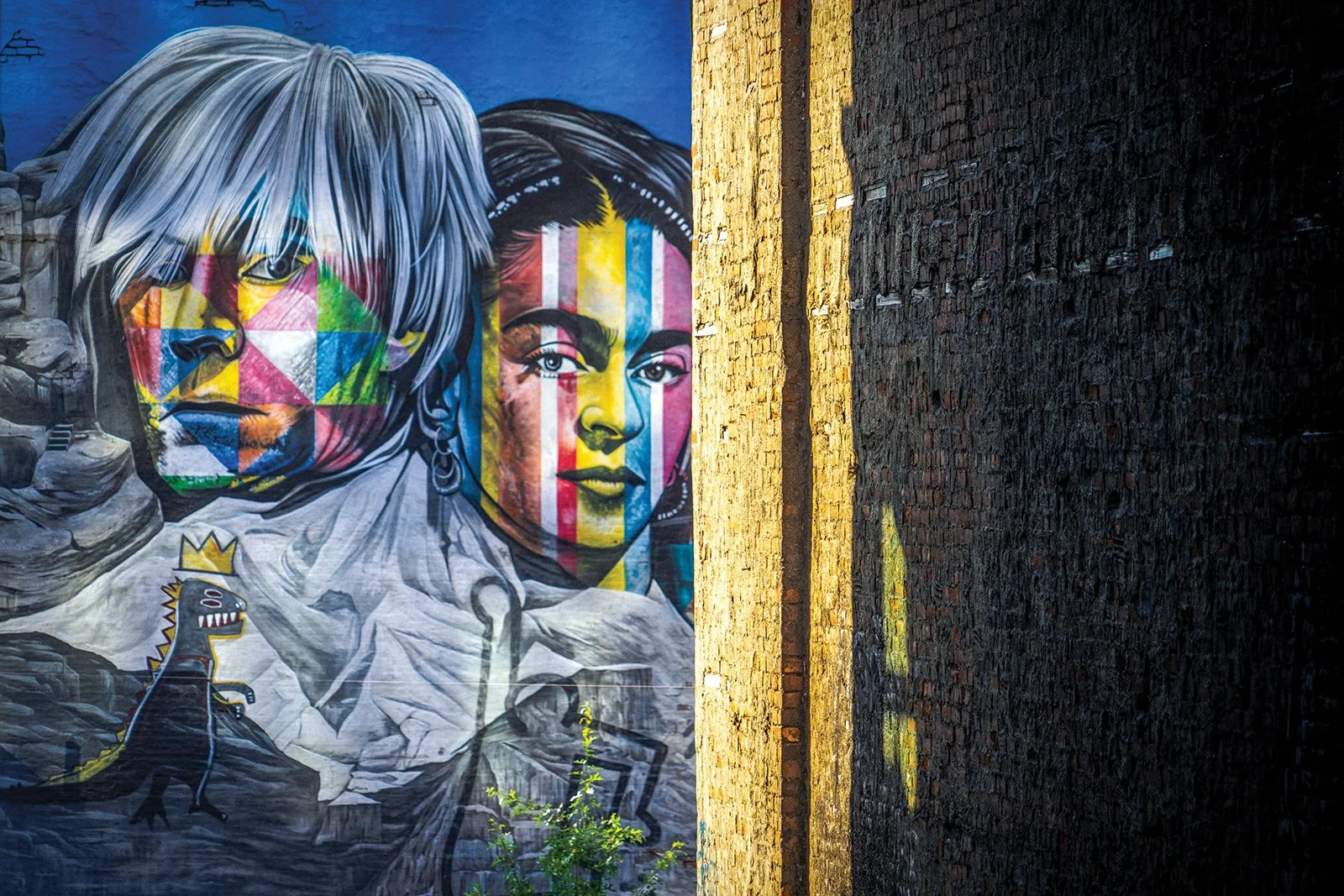 Colorful mural on a brick wall featuring two portraits: one of an older person with long, gray hair and serious expression, and a younger woman with dark hair and a confident look, with geometric and rainbow stripes over their faces, and a small cart