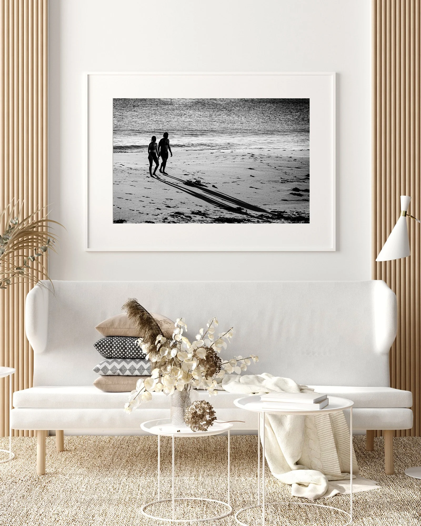 A living room with a white sofa, decorative pillows, a vase with dried flowers, and a wall art print of a black-and-white photo of a couple walking on the beach.