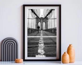 Black and white photo of a person walking on a bridge with suspension cables and towers, displayed in a black frame on a wall.