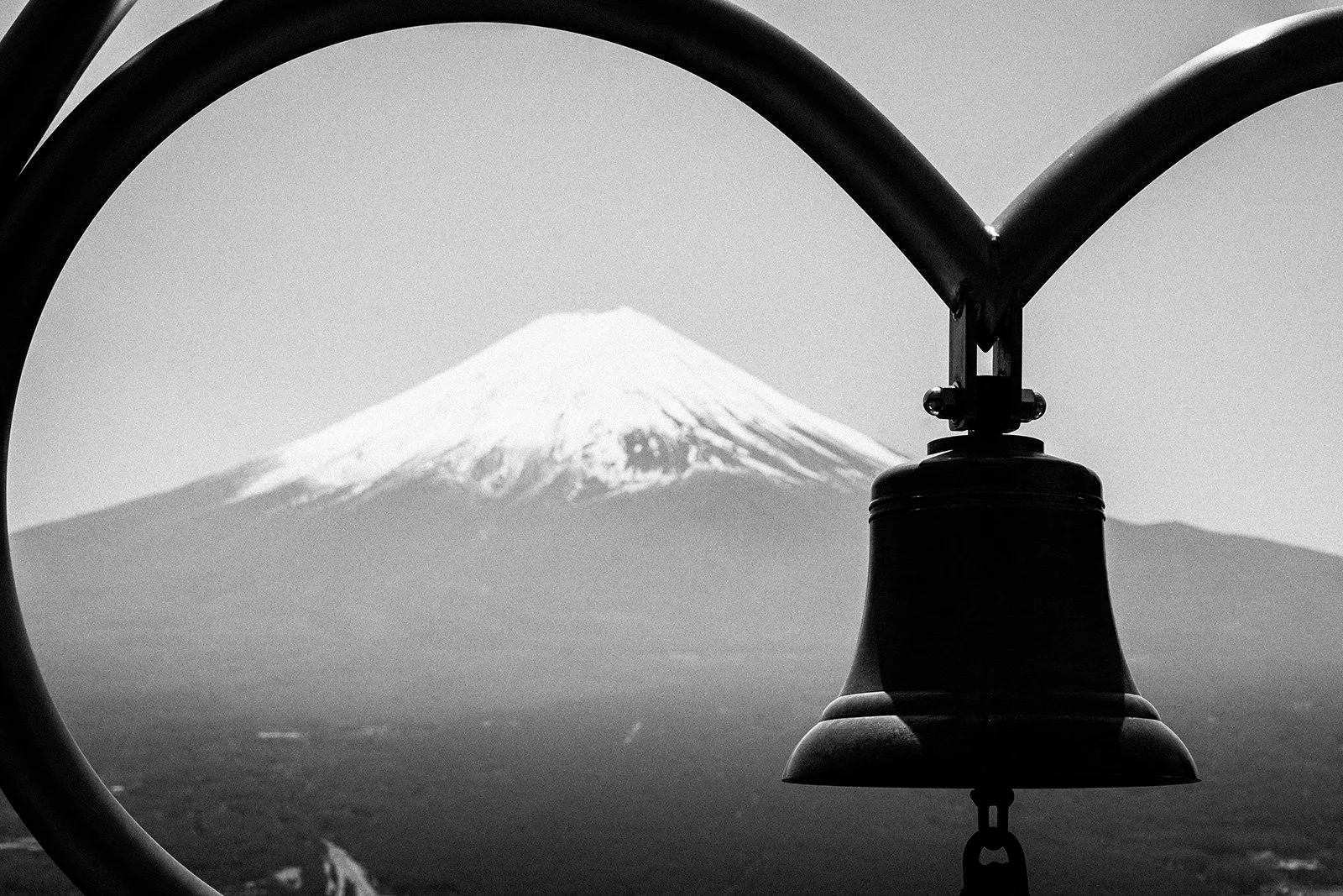 Black and white photograph of snow-capped Mount Fuji seen through a circular metal frame with a hanging bell in the foreground.