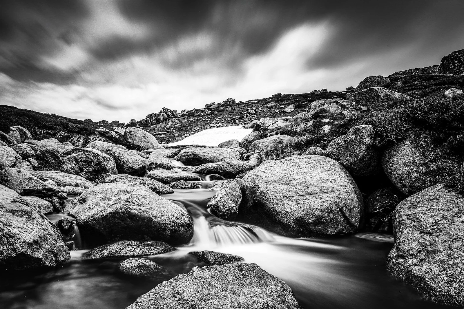 Black and white landscape photo of a rocky mountain stream with large boulders and cloudy sky.