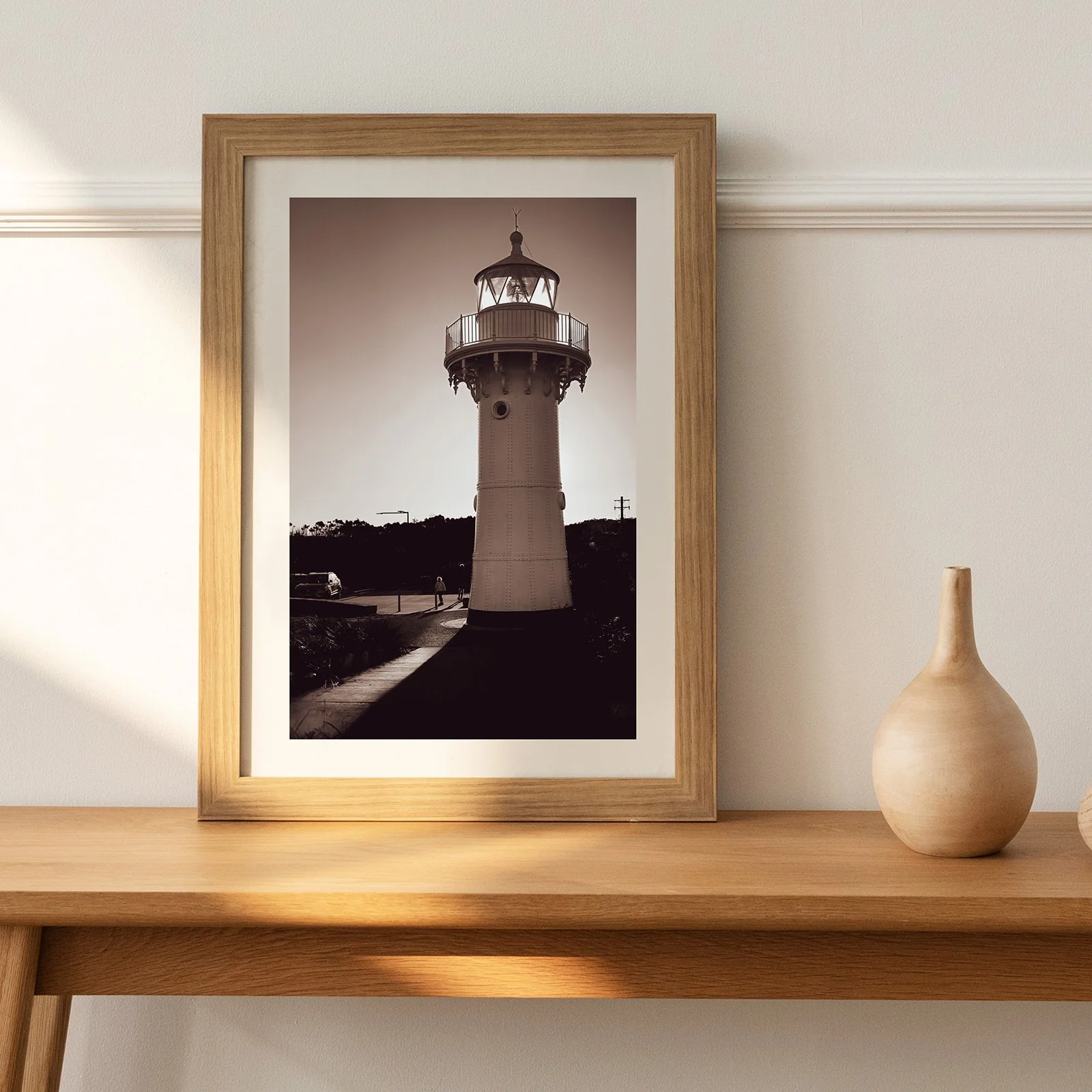 Framed black and white photograph of a lighthouse on a wooden shelf with a beige ceramic vase beside it, in a minimalist interior setting.