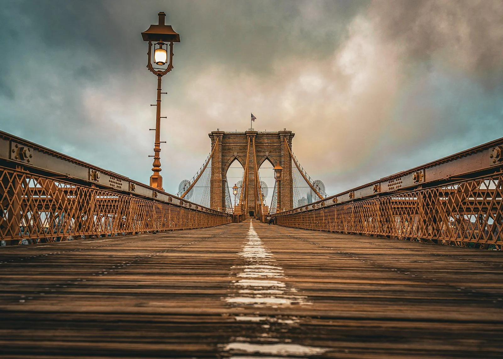 Low-angle view of Brooklyn Bridge with a cloudy sky in the background, showcasing its wooden walkway, metal railings, and stone towers.
