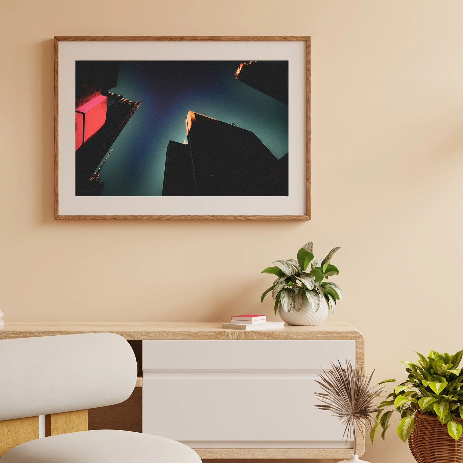 A room with a light-colored wall, a wooden framed photograph of a skyscraper shot from below, a white cabinet with a potted plant, books, and a white chair with a wooden base, and another potted plant in a brown wicker basket.