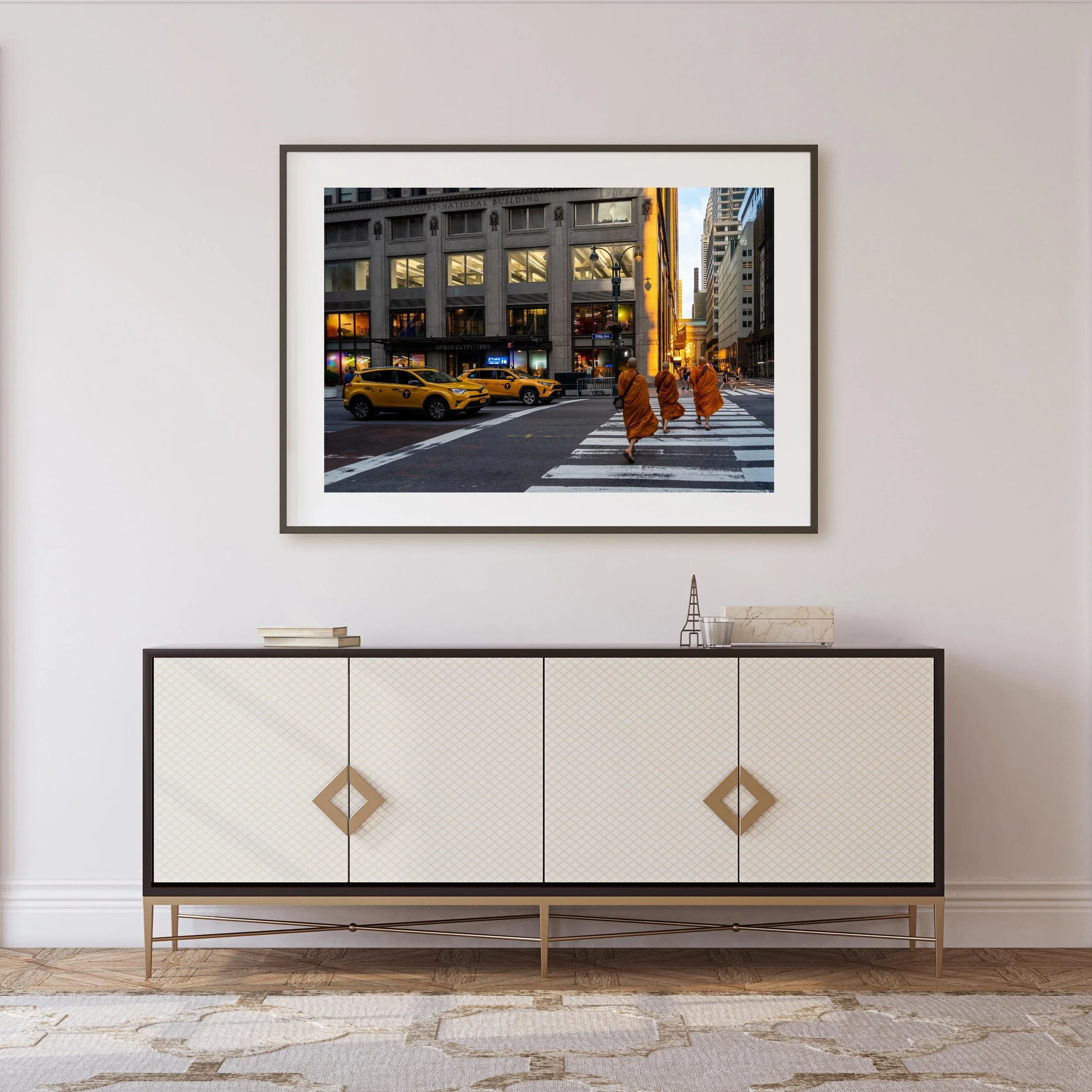 Photograph of a city street scene at sunset, with four monks walking across a crosswalk and yellow taxis parked along the street, viewed from inside a building with a white wall and a modern console table underneath the picture.