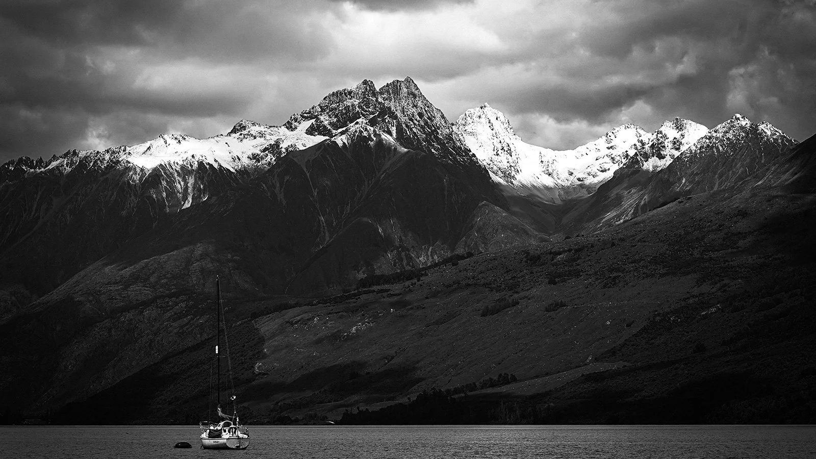 Black and white landscape of a mountain range with snow-capped peaks, a dark cloudy sky, and a sailboat on calm water in the foreground.