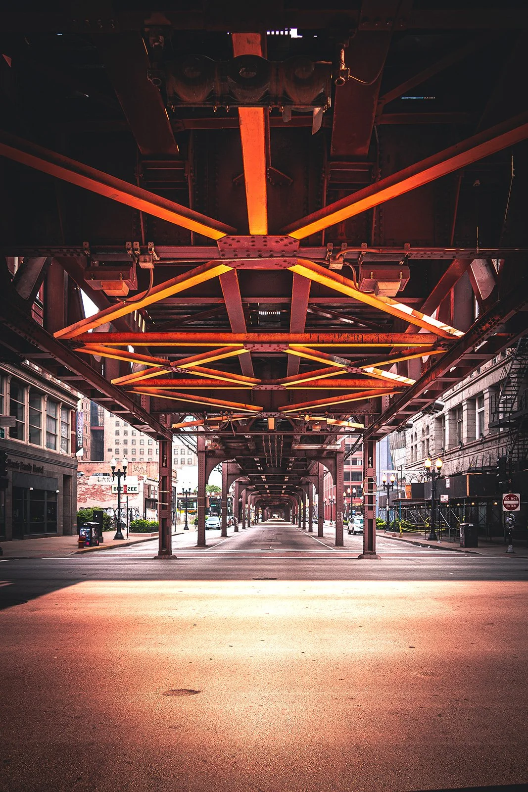 View of an urban street scene under an elevated train track with red metal supports and orange lighting, empty road, and surrounding buildings.