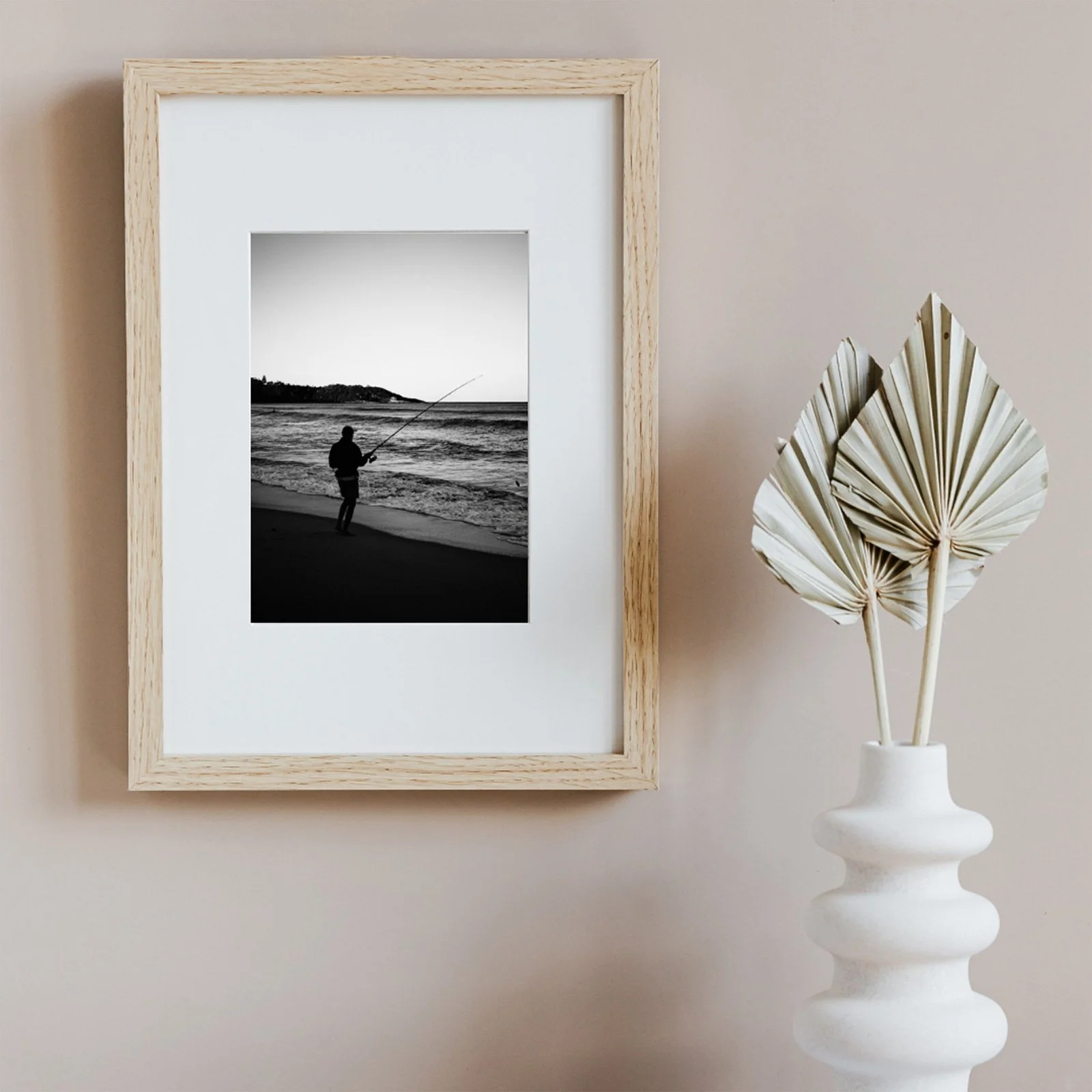 Black and white photograph of a person fishing on a beach, framed on a wall next to a white vase with dried palm leaves.