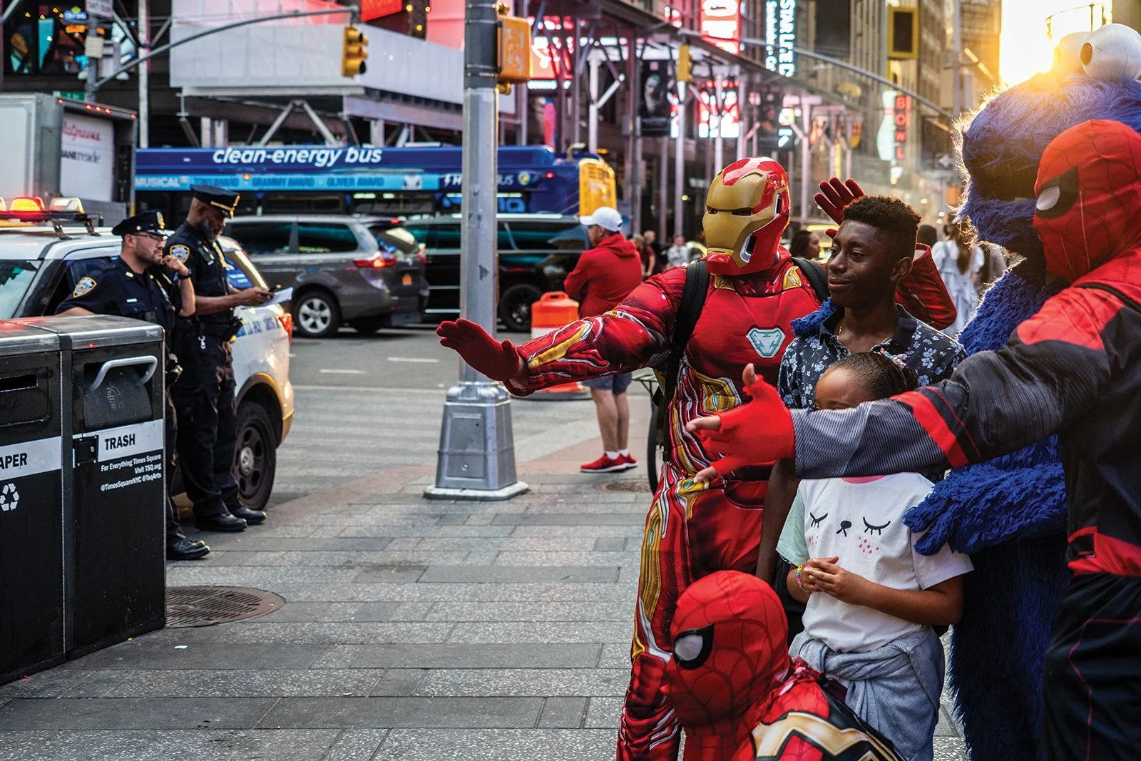 People dressed in superhero costumes interacting with children on a city street, with police officers and vehicles in the background.