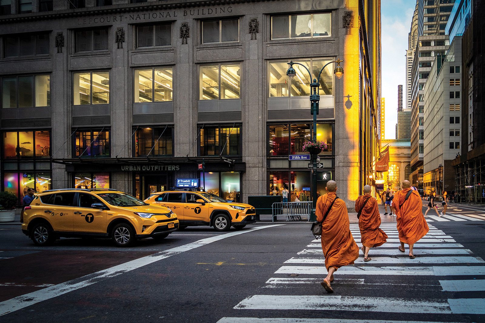 Four Buddhist monks walking across a city crosswalk at sunset, with yellow taxis and tall buildings in the background.