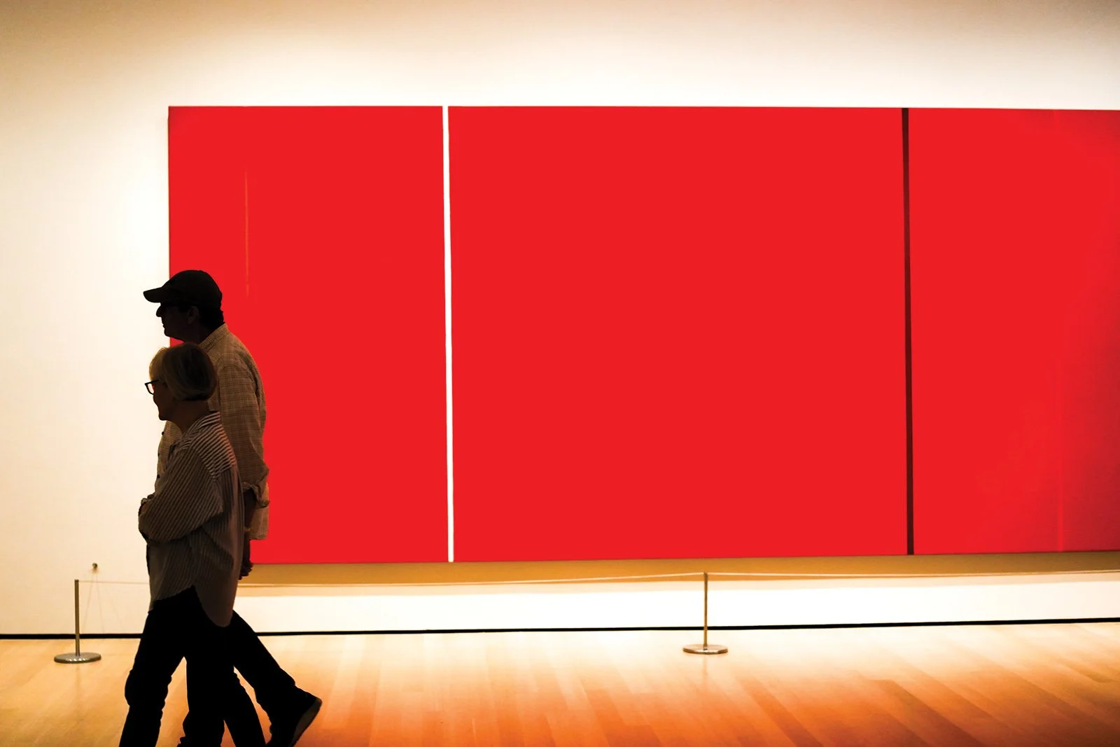 Silhouettes of three people walking past a large red artwork in a gallery.