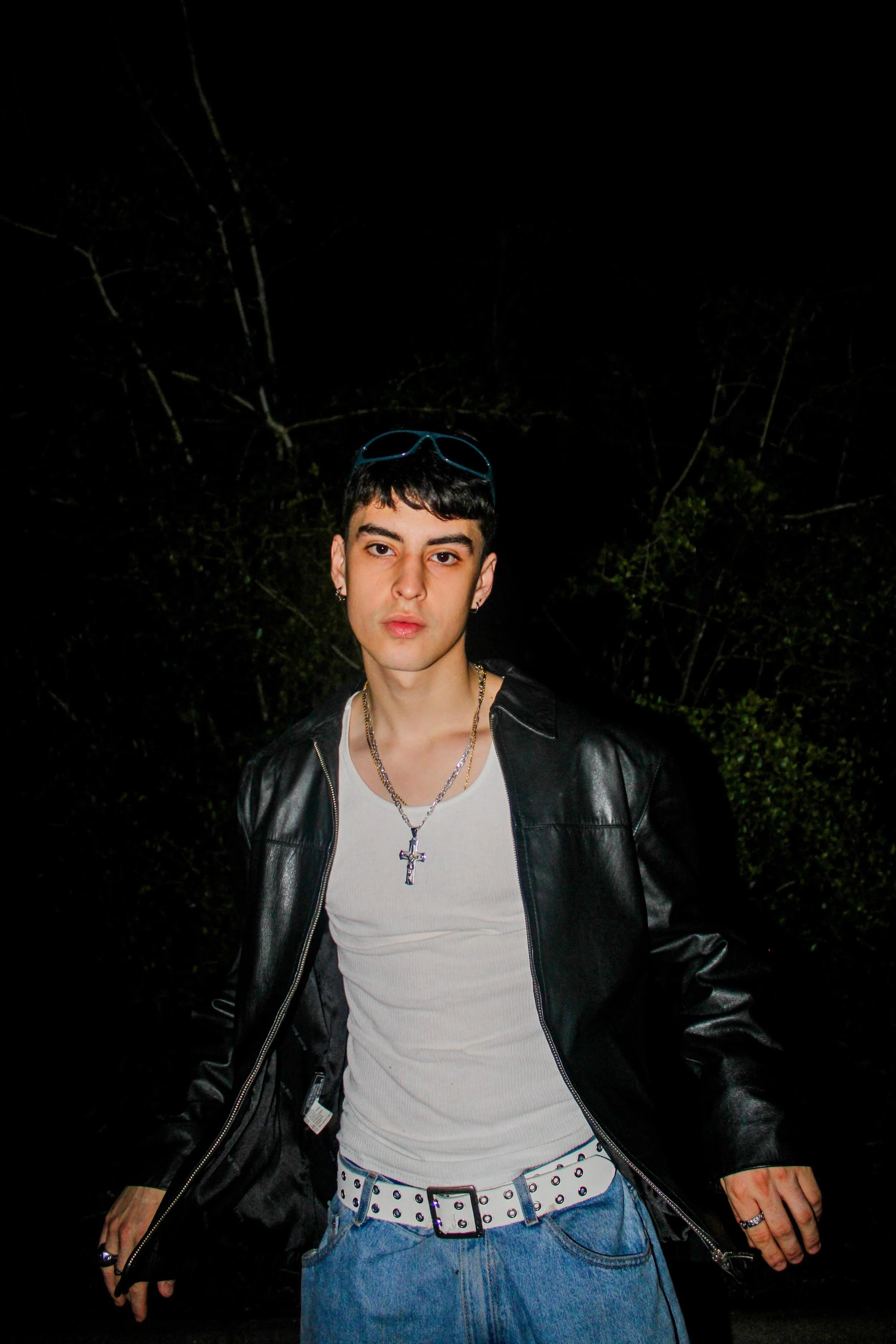 Young man with short dark hair, wearing sunglasses on his head, a cross necklace, a white t-shirt, a black leather jacket, and blue jeans with a studded belt, standing outdoors at night.