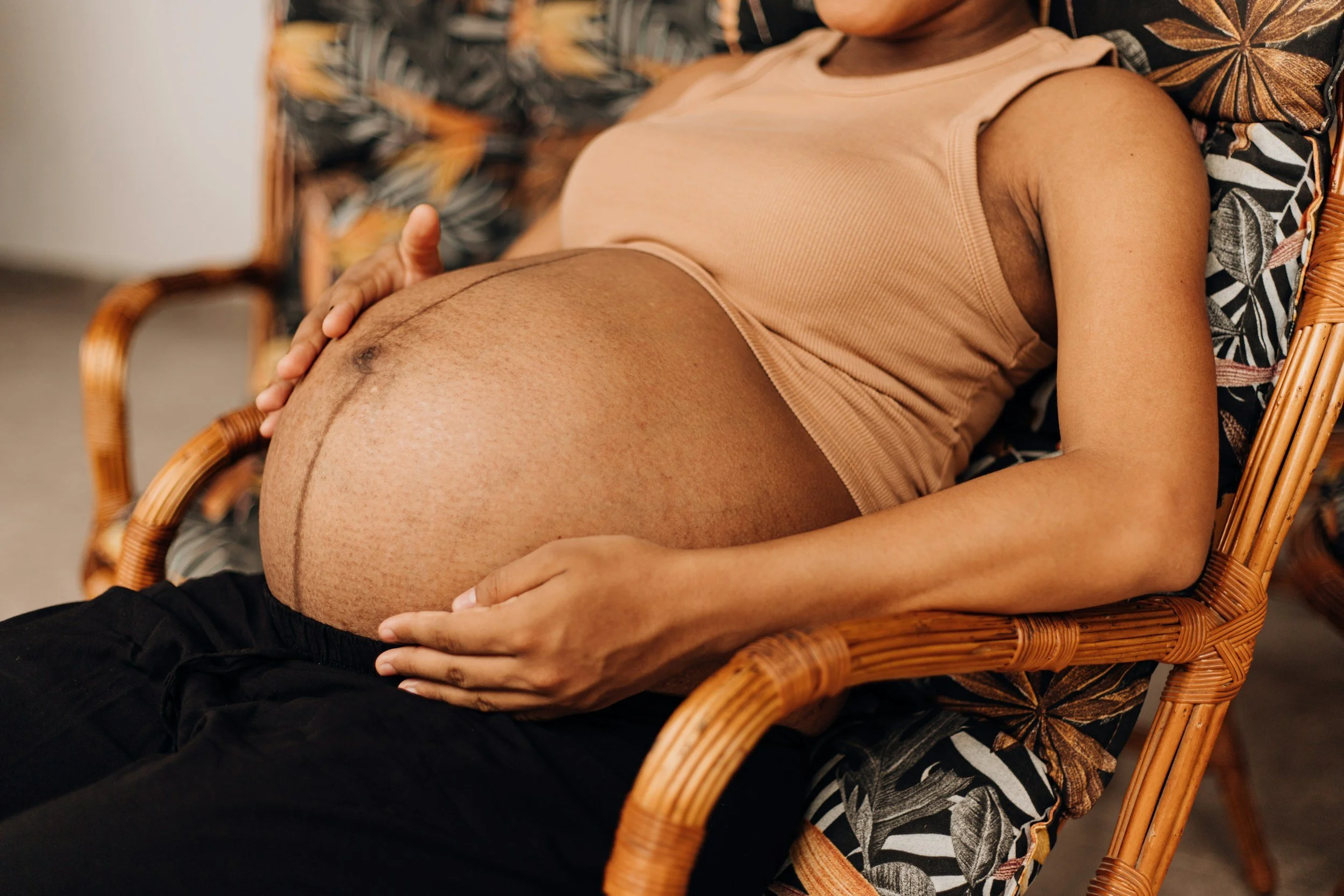 A person with dark skin, wearing a beige sleeveless top, resting on a patterned chair with a rounded rattan armrest.