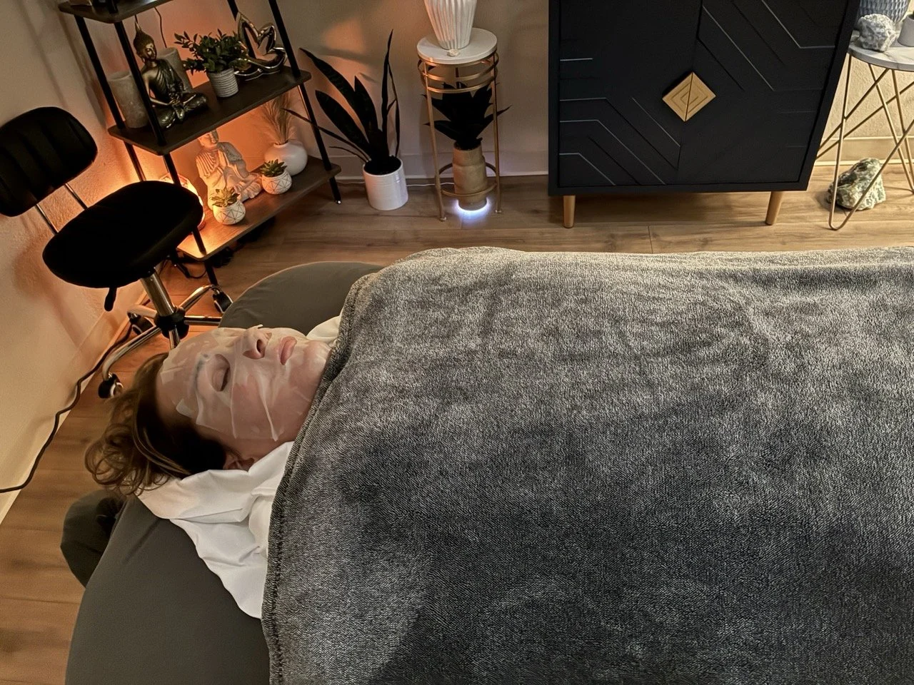 Person lying on massage table with face mask and blanket in a cozy, well-decorated room with plants, shelves, and elegant furniture.