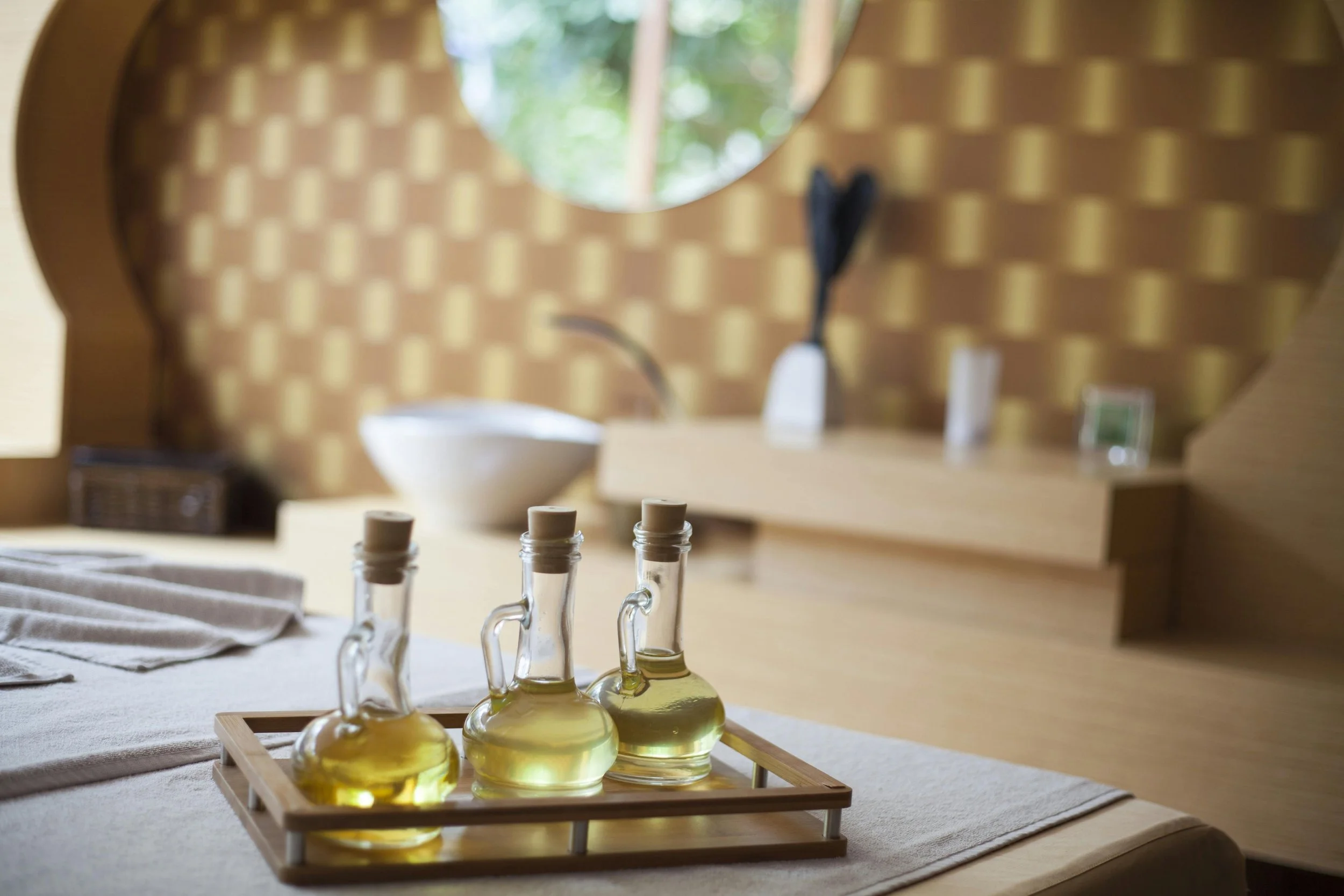 Glass bottles filled with yellow liquid arranged on a wooden tray on a beige cloth, with a blurred background of a room with a mirror and patterned wall.