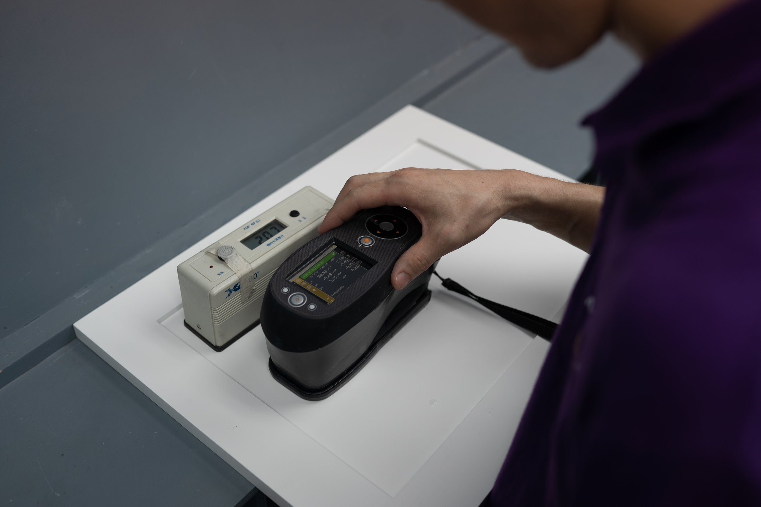 A person in a purple shirt is holding a handheld device, likely a Geiger counter, over a white surface with an electronic device connected to a cord. The electronic device displays readings on a small screen, and the setup appears to be in a laboratory or testing environment.