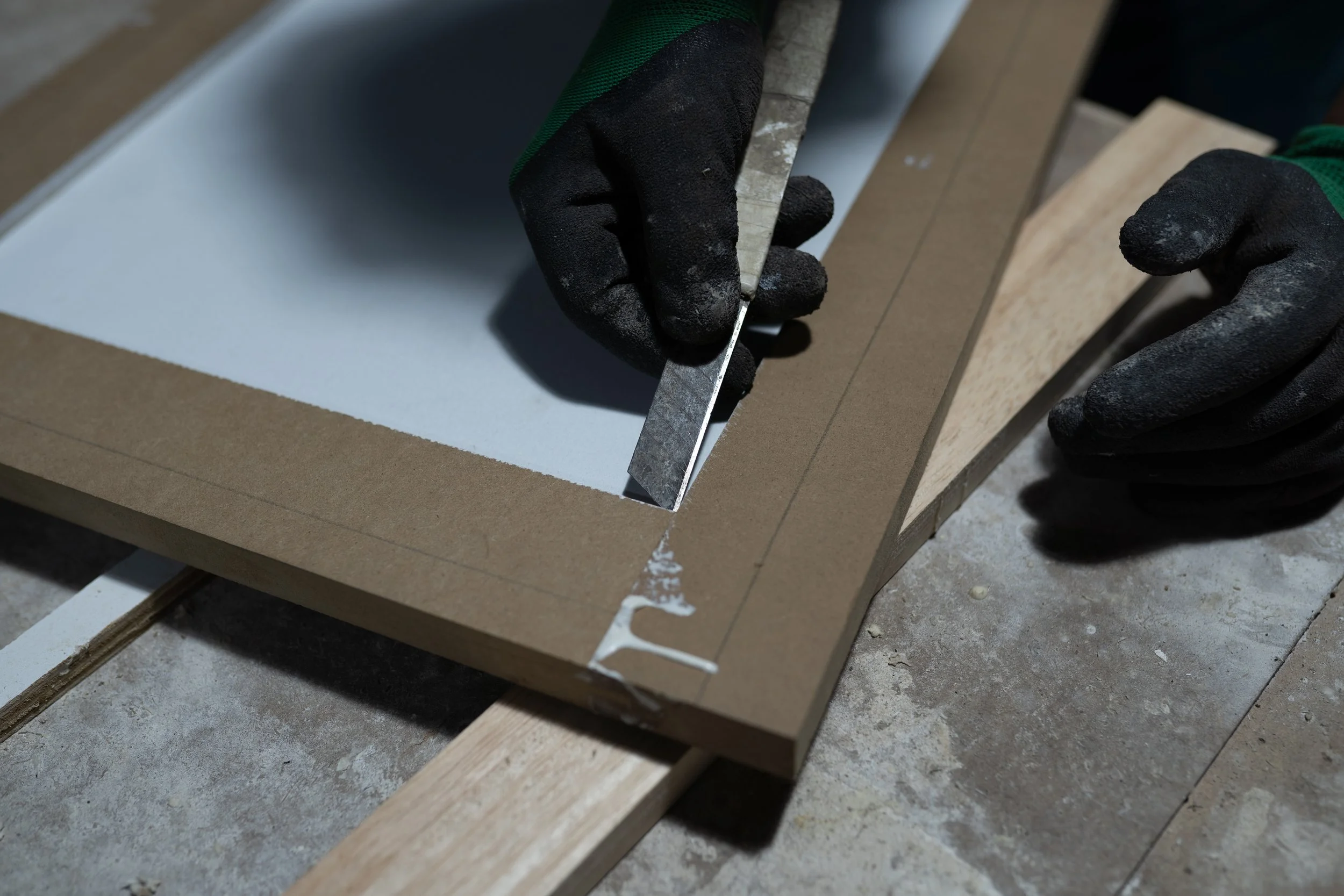 Close-up of hands wearing black gloves cutting a piece of wood with a metal ruler in a woodworking shop.