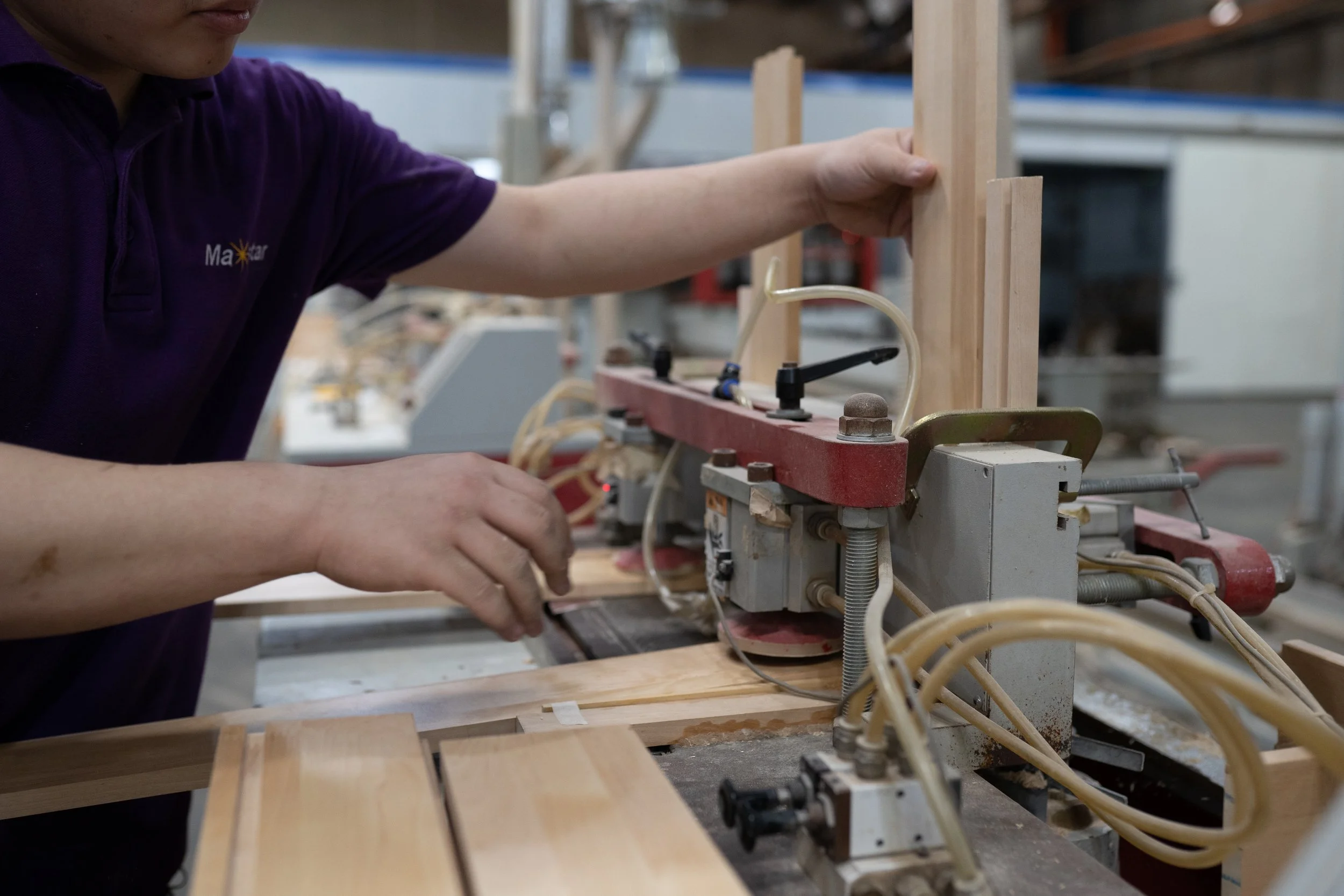 A person in a purple shirt operates a woodworking machine, shaping a piece of wood in a workshop.