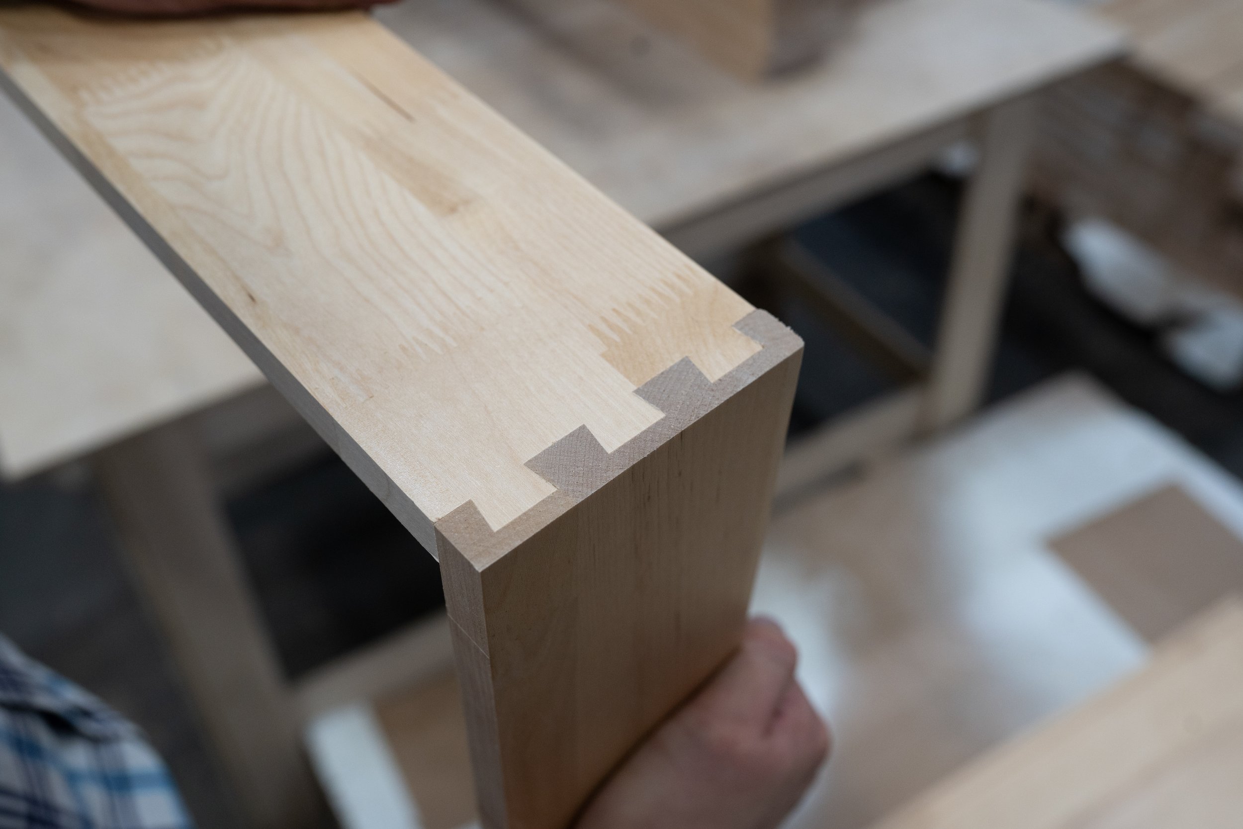 Close-up of a light-colored wooden piece with visible grain and a dovetail joint at the corner, held above a workbench.