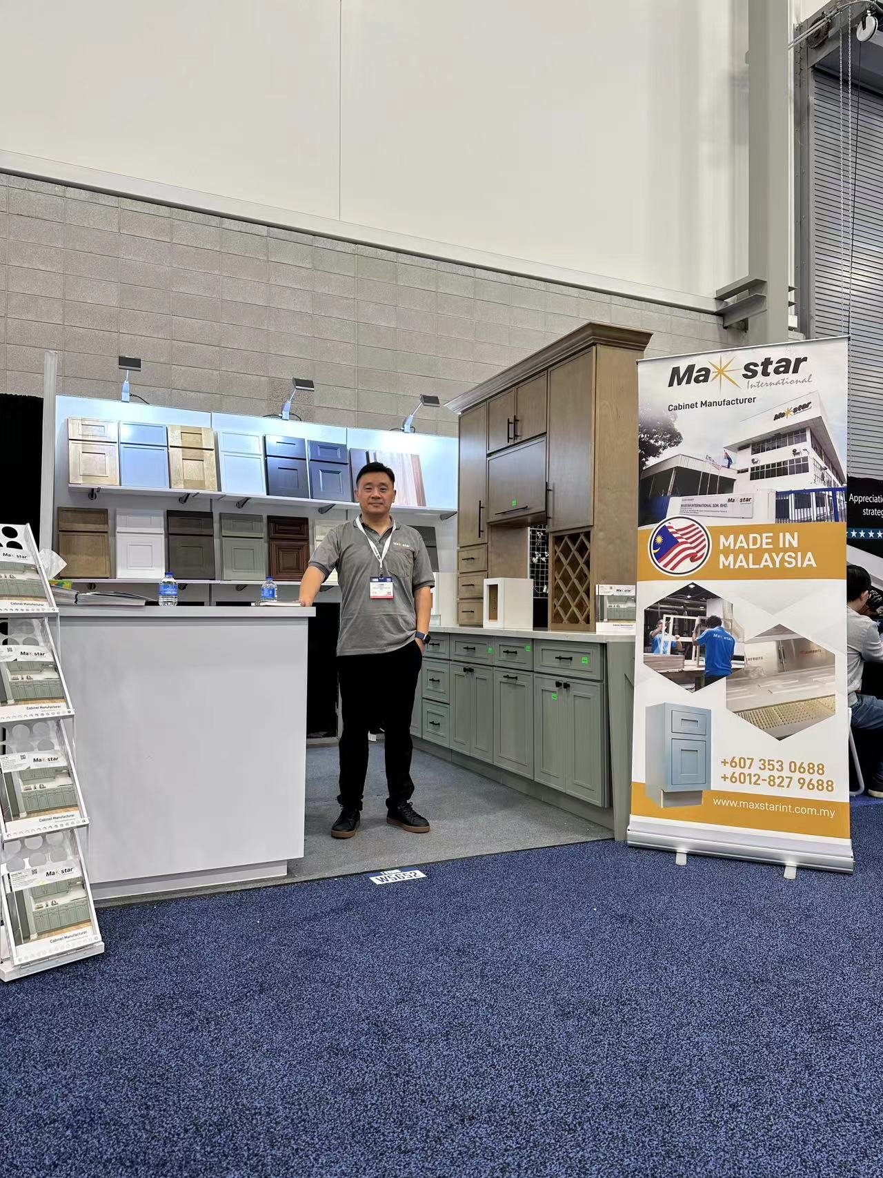 A man standing inside an exhibition booth displaying various cabinets and kitchen furniture. There is a banner next to him with the logo 'Max star', indicating they are a cabinet manufacturer made in Malaysia. The booth is set up in a convention center with a blue carpeted floor.