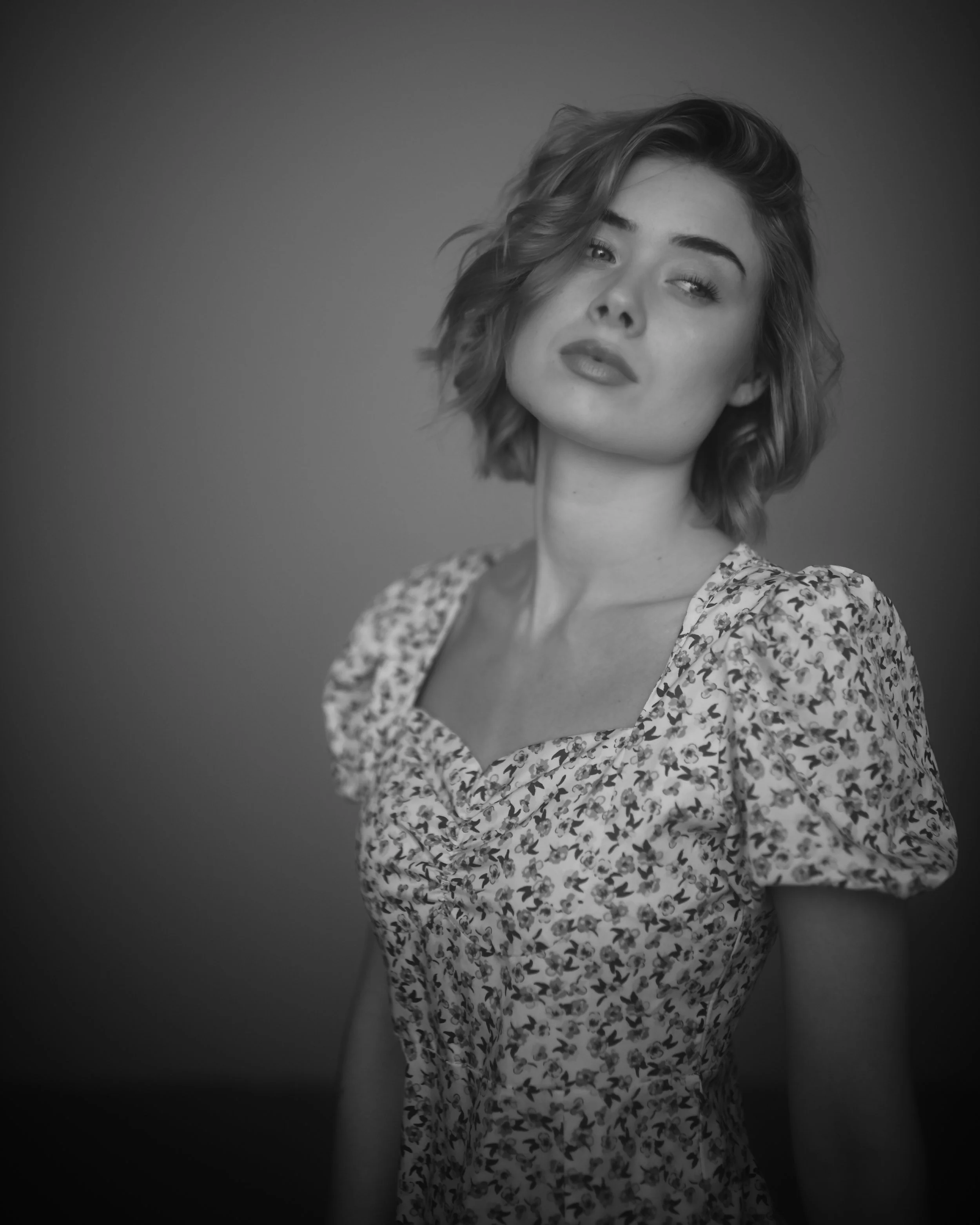 Schwarz-weiß Portrait eines jungen Mädchens mit lockigem, mittellangem Haar, das leicht nach oben frisiert ist. Sie trägt ein Kleid mit Herzmustern und hat einen lockeren, nachlässigen Ausdruck.