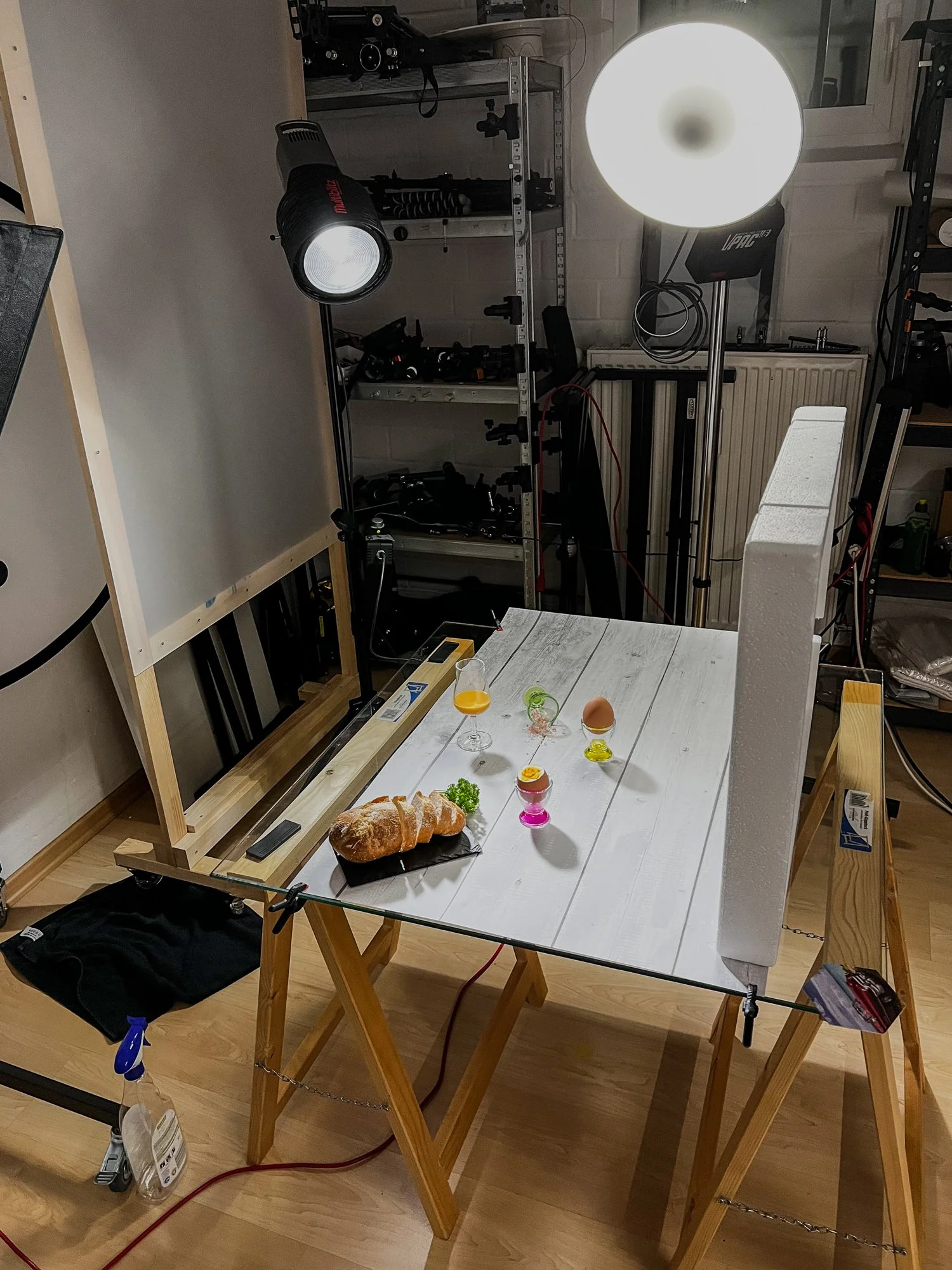 Auf einem Tisch stehen Lebensmittel, darunter ein Brot, Eier, Gläser mit Getränken und Gemüse. Im Hintergrund sind Fotogeräte und Equipment für eine Fotosession sichtbar, darunter Lichter und Reflexoren.