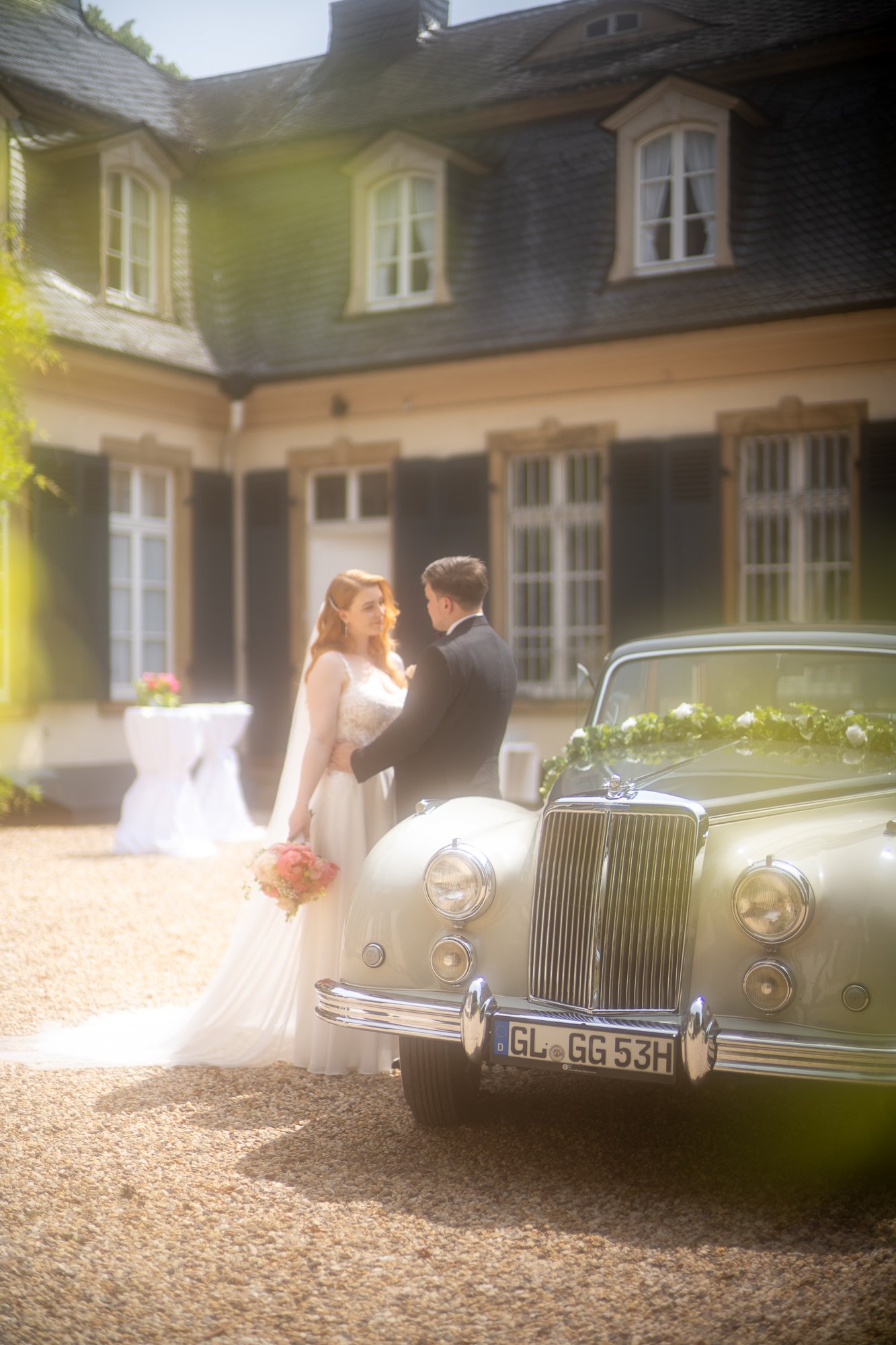 Brautpaar steht vor einem klassischen schwarzen Auto vor einem historischen Haus mit dunklen und weißen Fensterrahmen, während sie sich anschauen. Es ist eine Hochzeitsszene im Freien.