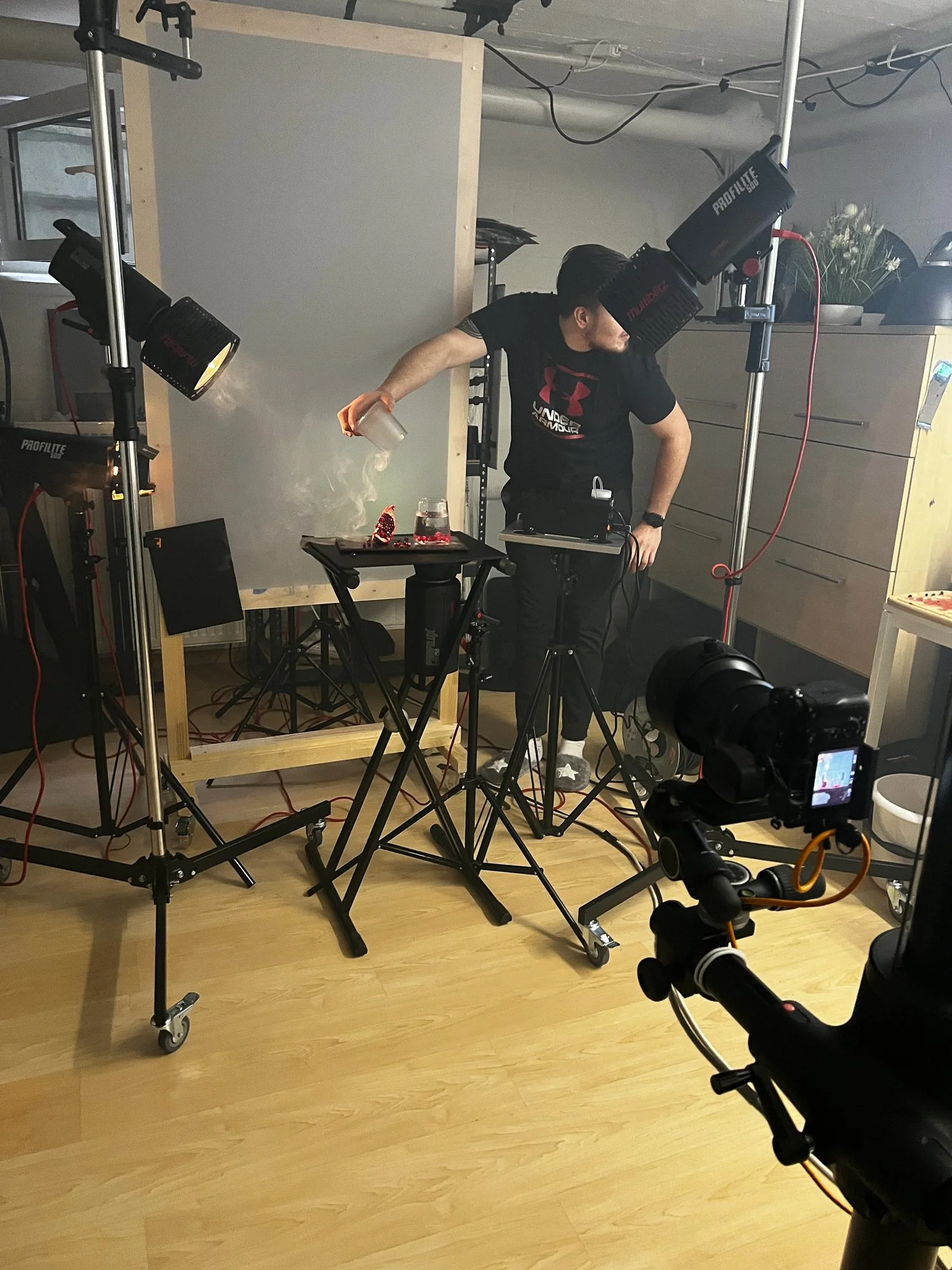Fotostudio mit Beleuchtungsausrüstung, Kamera, Studiotisch, auf dem ein Mann mit schwarzen Haaren und einem schwarzen T-Shirt Getränk spuckt, während eine andere Kamera aufnimmt.