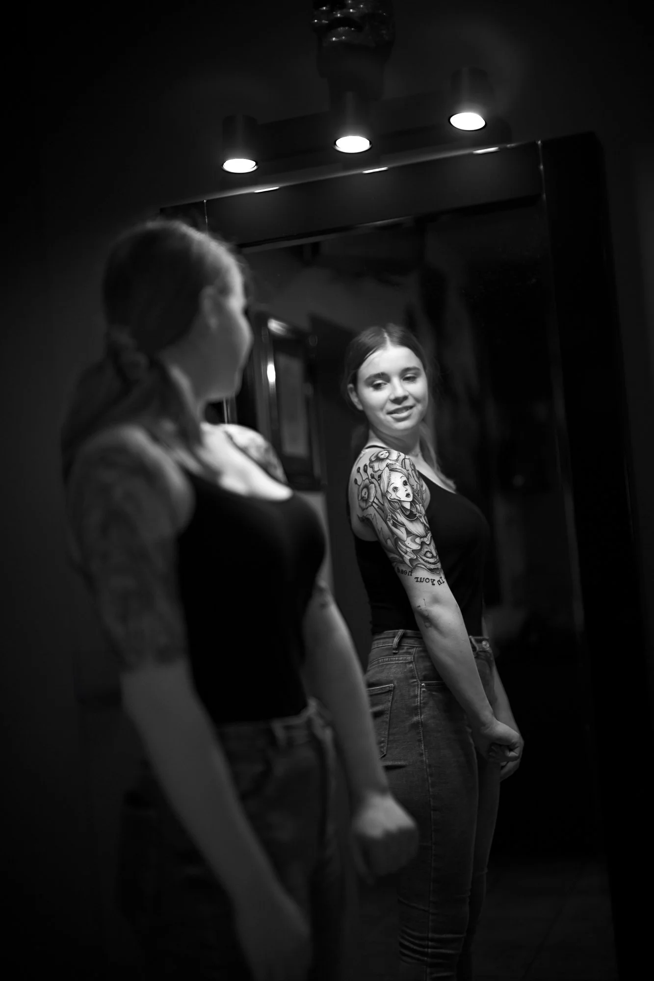Eine junge Frau mit Tattoos auf dem Arm, die vor einem Spiegel steht und in die Kamera lächelt, während sie sich im Spiegel betrachtet.