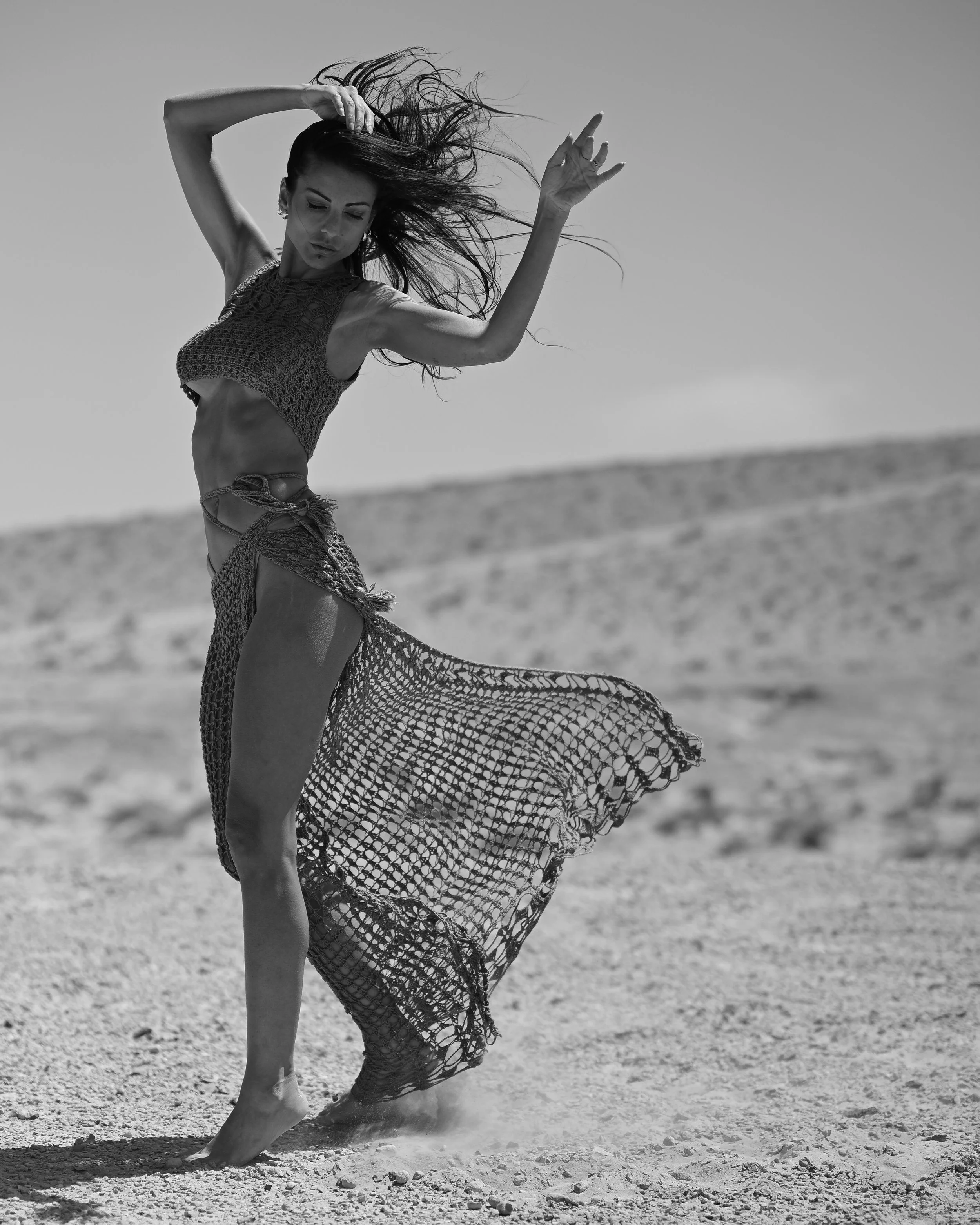Schwarz-Weiß-Foto einer Frau, die auf einem Strand tanzt, mit wehender Kleidung und nassen Haaren, bedeckt den Boden mit Staub und Sand, im Hintergrund Himmel und Meer.