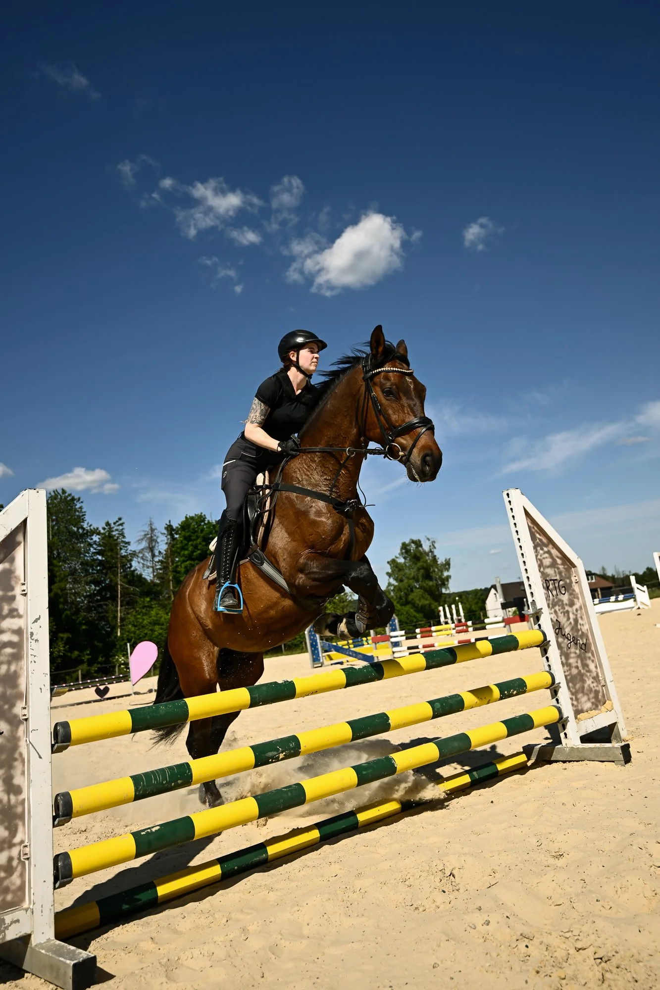Reiter springt mit seinem Pferd über einen Hinderniss in einem Springreitwettbewerb im Freien bei sonnigem Wetter, mit blauer Himmel und weißen Wolken, in einem Reitstall oder -platz.
