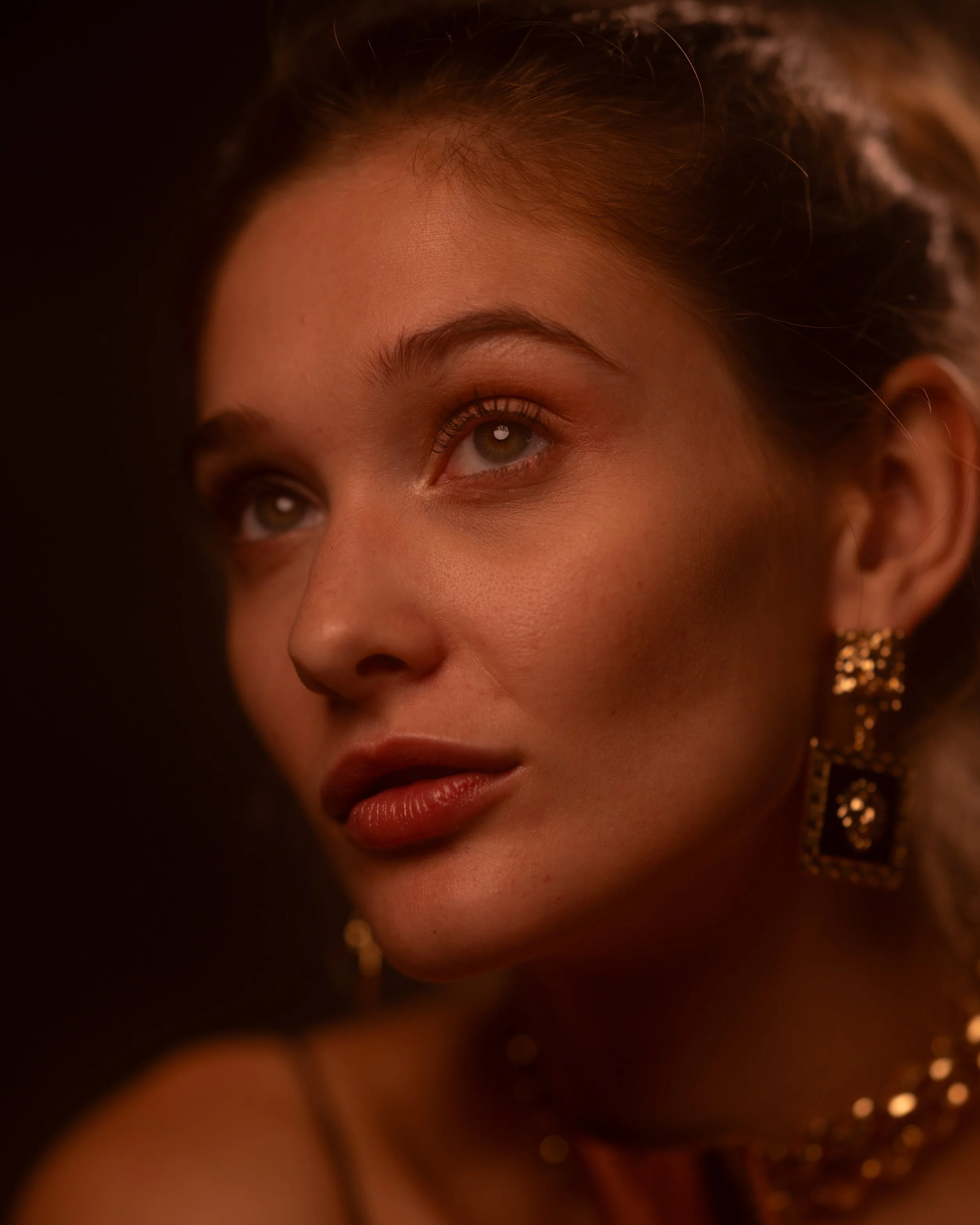 Nahaufnahme eines FrauenGesichtes mit braunen Augen, geschminkten Lippen, goldener Ohrringe, dunklem seidigem Haar und einem warmen, weichen Licht