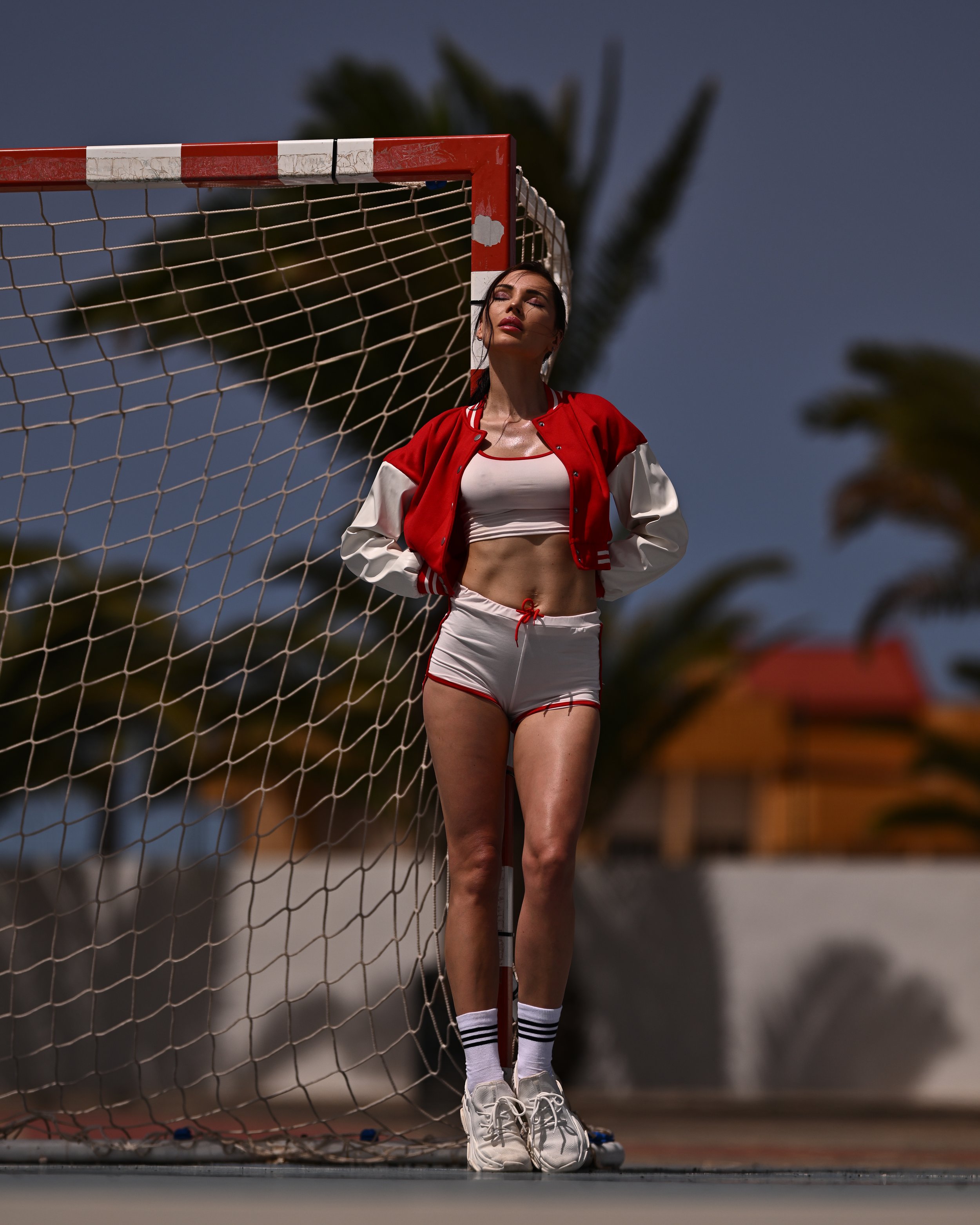 Eine junge Frau steht im Sportoutfit an einem Fußballtor, trägt weiße Turnschuhe, Socken mit schwarzen Streifen, Shorts und eine rote Jacke, während sie in der Sonne steht, mit Palmen im Hintergrund.